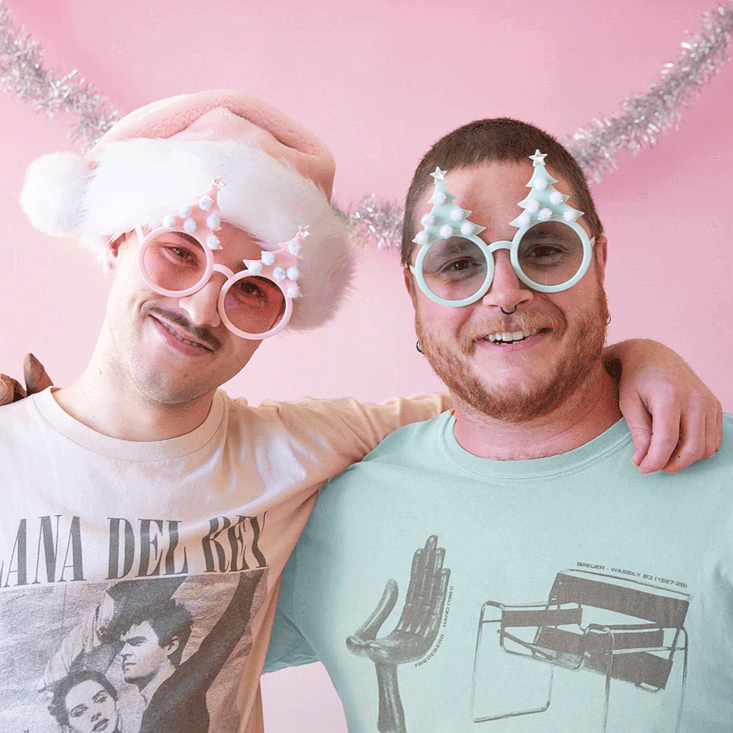 On a pink background is two models wearing the christmas tree holiday glasses. One is wearing he pastel pink version and the other is wearing the pastel mint shade. Each pair of glasses has round lenses and six white pom poms on each tree shape.