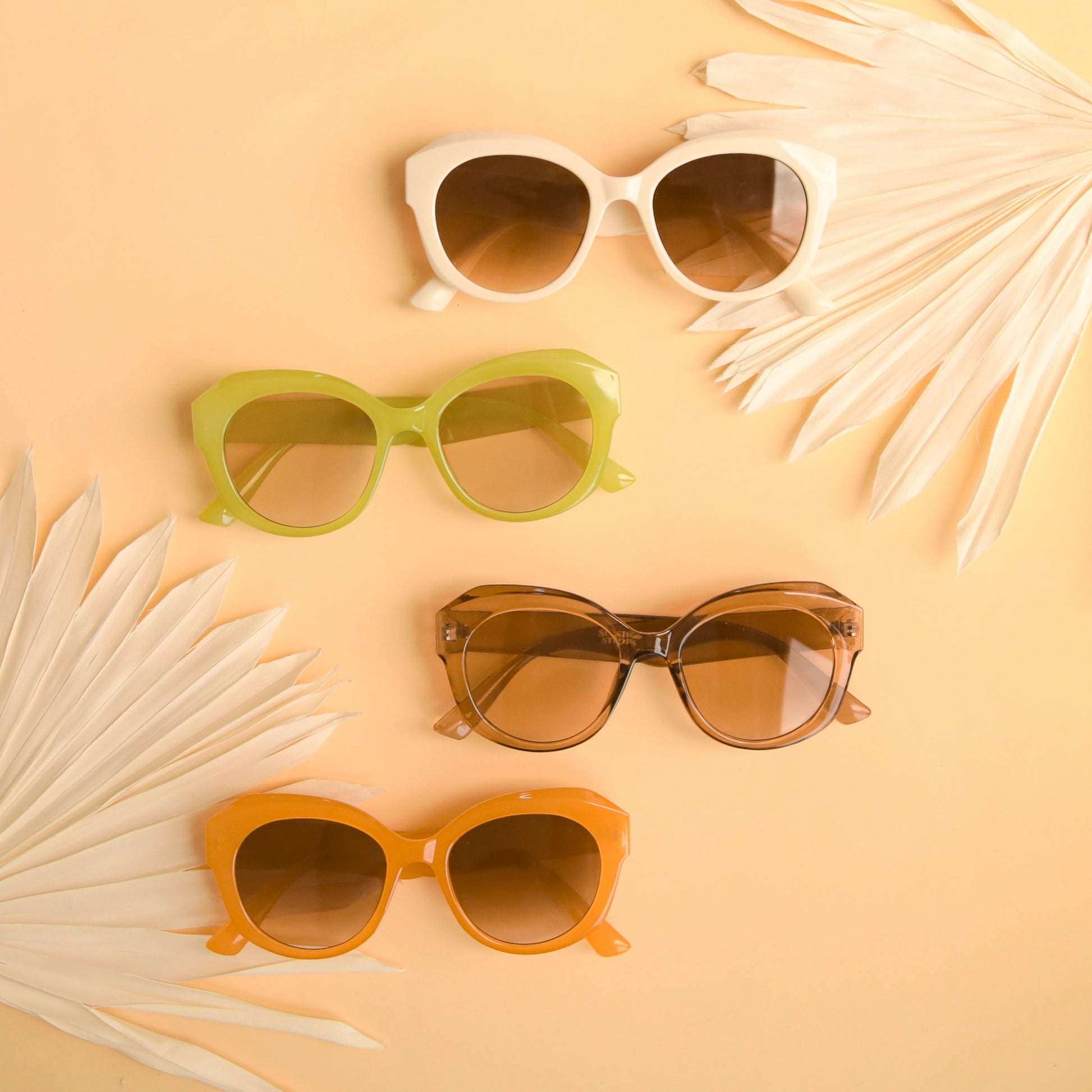 On a peachy background is a group photo of the different color options that the Donna Sunglasses come in. There is a white pair, a green, a light brown and an orange.