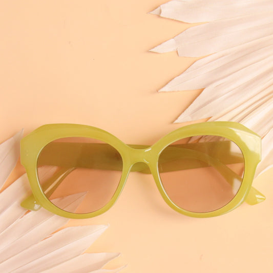 On a peachy background is a round pair of light green sunglasses with a light brown lens. 
