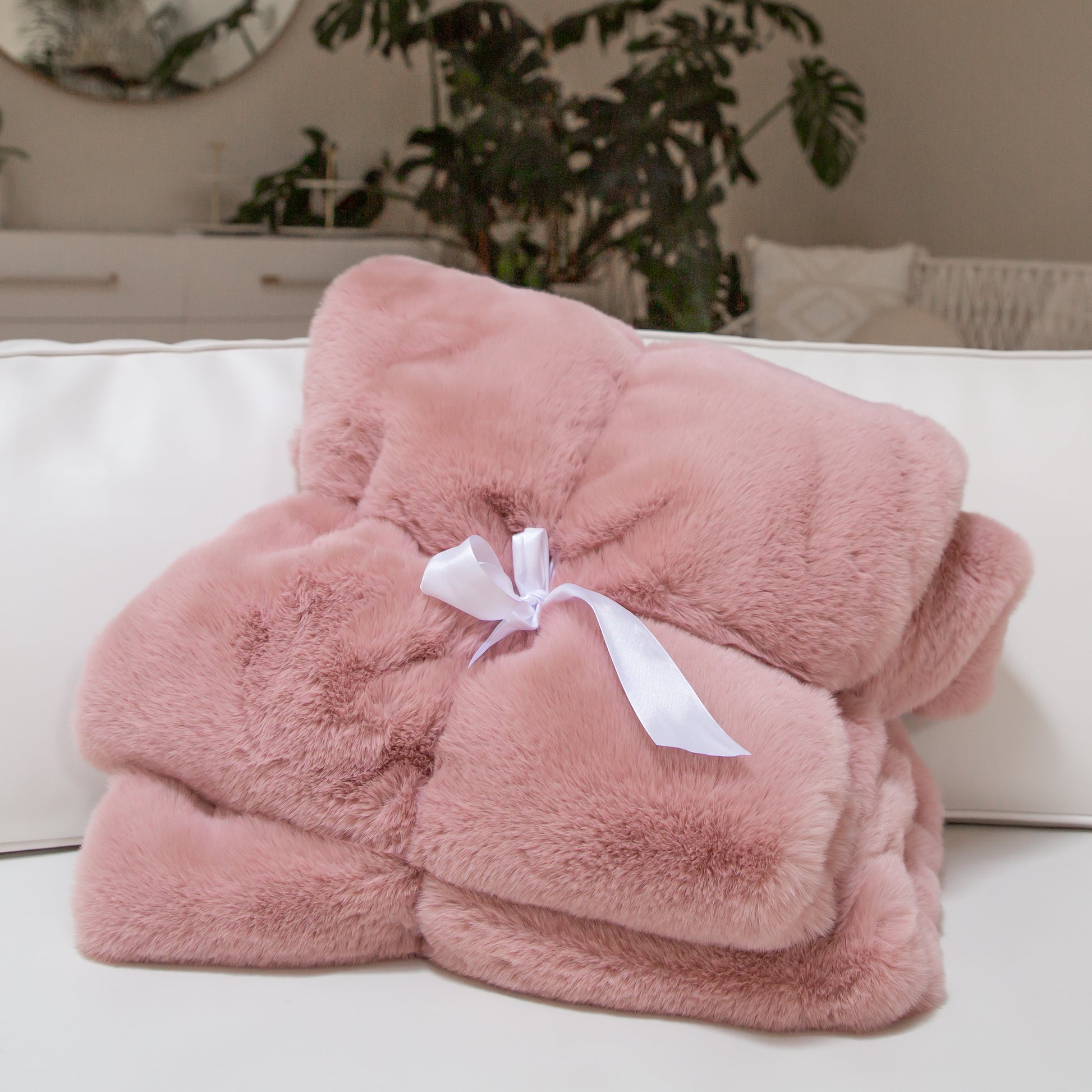 On a tan background is a dusty rose faux fur throw blanket folded with a white satin bow in the center. 