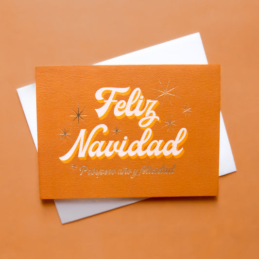 Burnt orange greeting card that reads 'Feliz Navidad' in white cursive lettering. Below reads 'prosper año y felicidad' in gold foil lettering. Gold foil stars twinkle behind the text. 