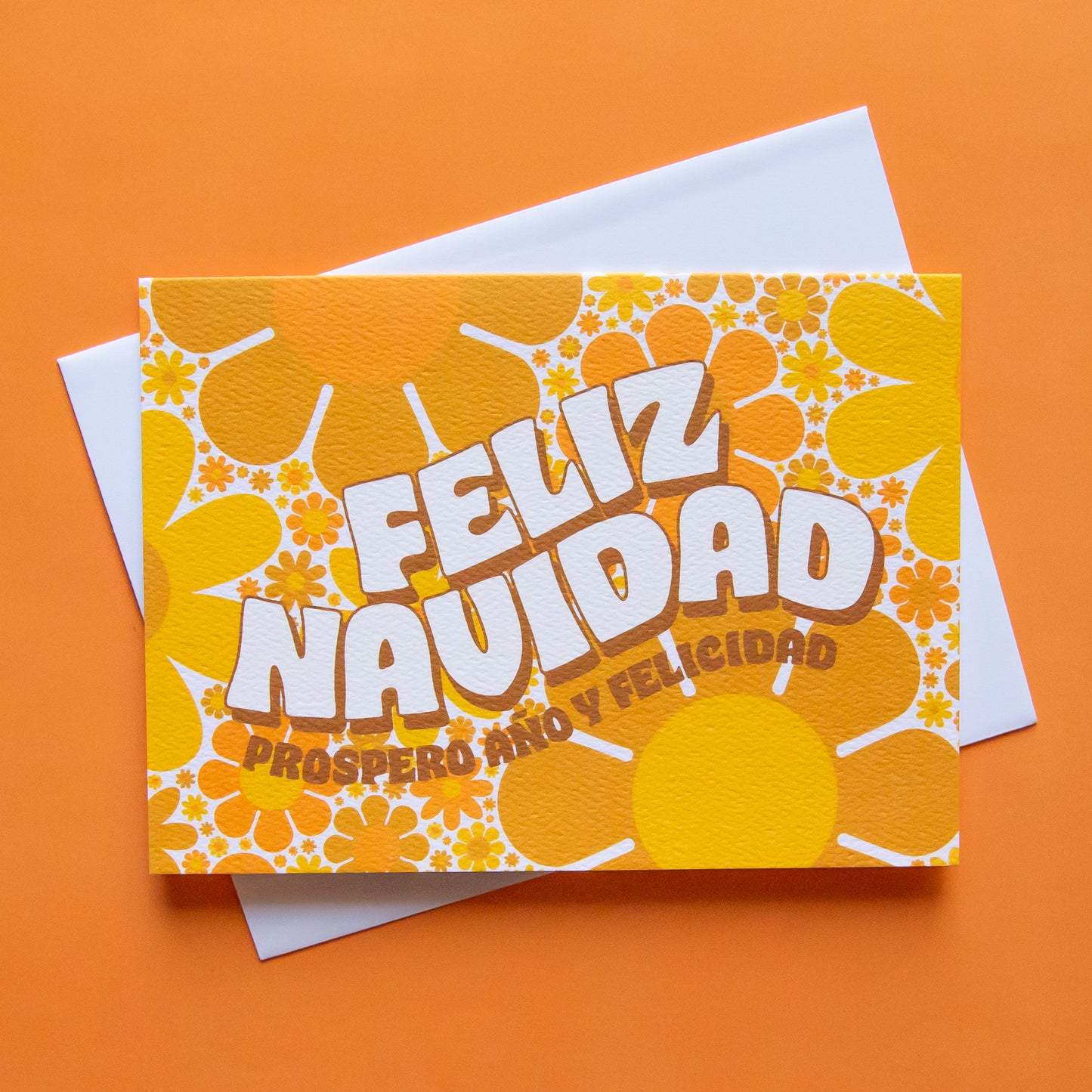 Greeting card filled with yellow and orange retro flower print. The card reads 'Feliz Navidad' in white curved bubble letters. Below reads 'prosper año y felicidad' in rust colored lettering. The card is accompanied by a solid white envelope. 