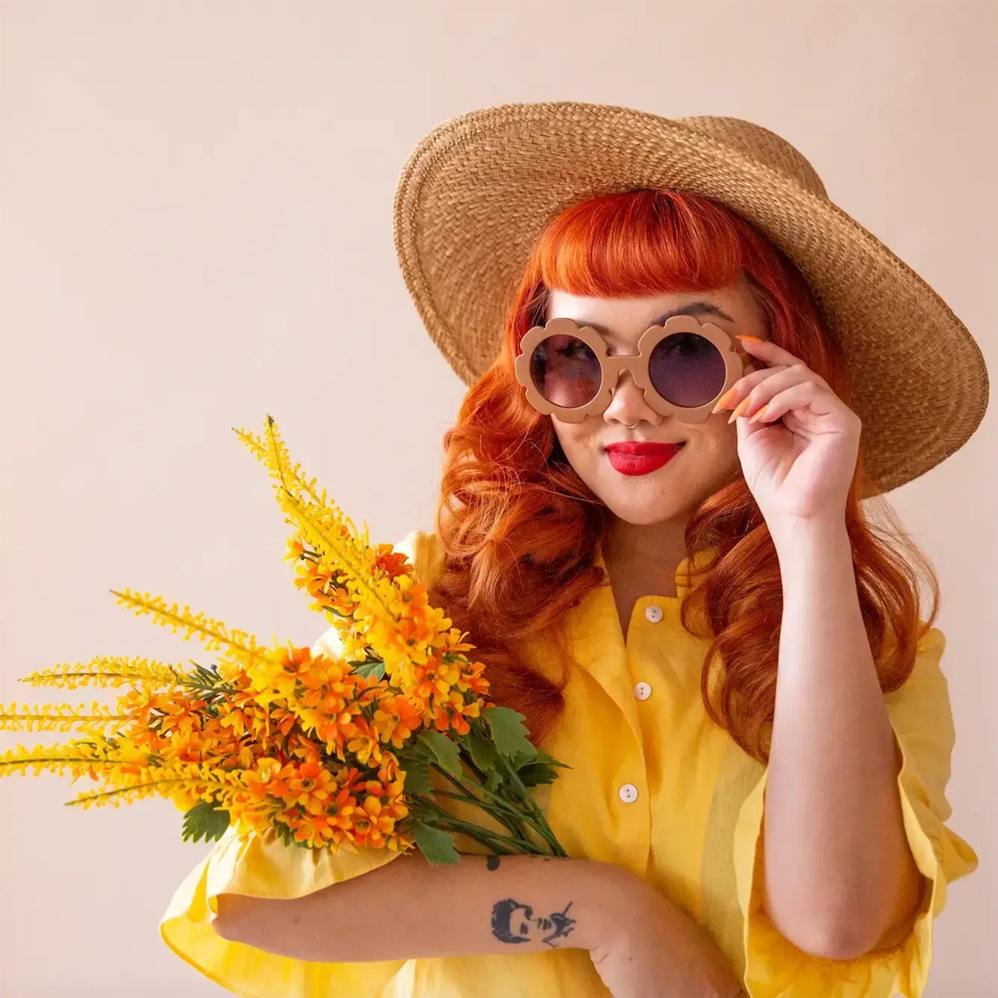 On an ivory background is a model wearing a pair of brown, flower shaped sunglasses and holding a bouquet of yellow flowers.