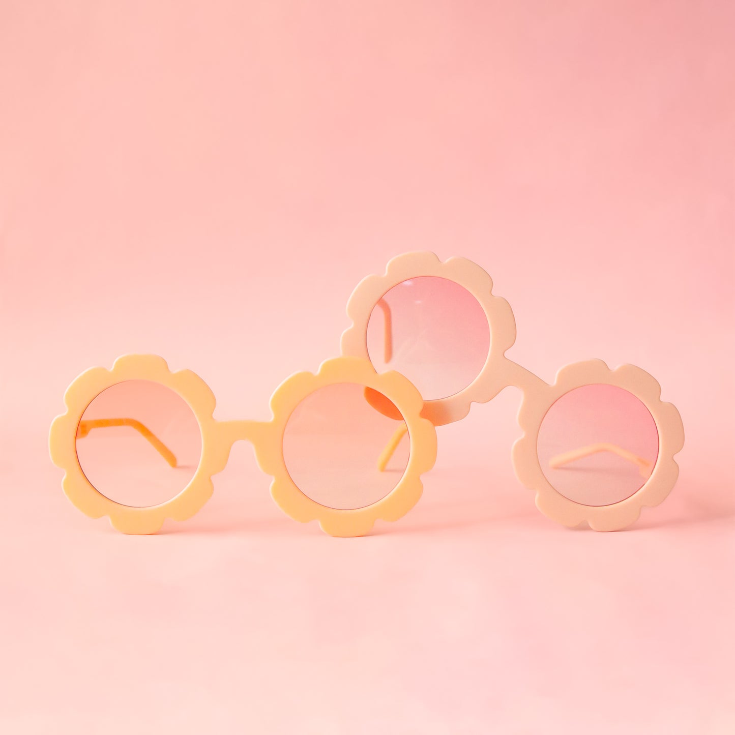 On a peachy background is a pair of flower shaped sunglasses with pink lenses and a apricot colored frame with the pink version of the flower sunglasses.