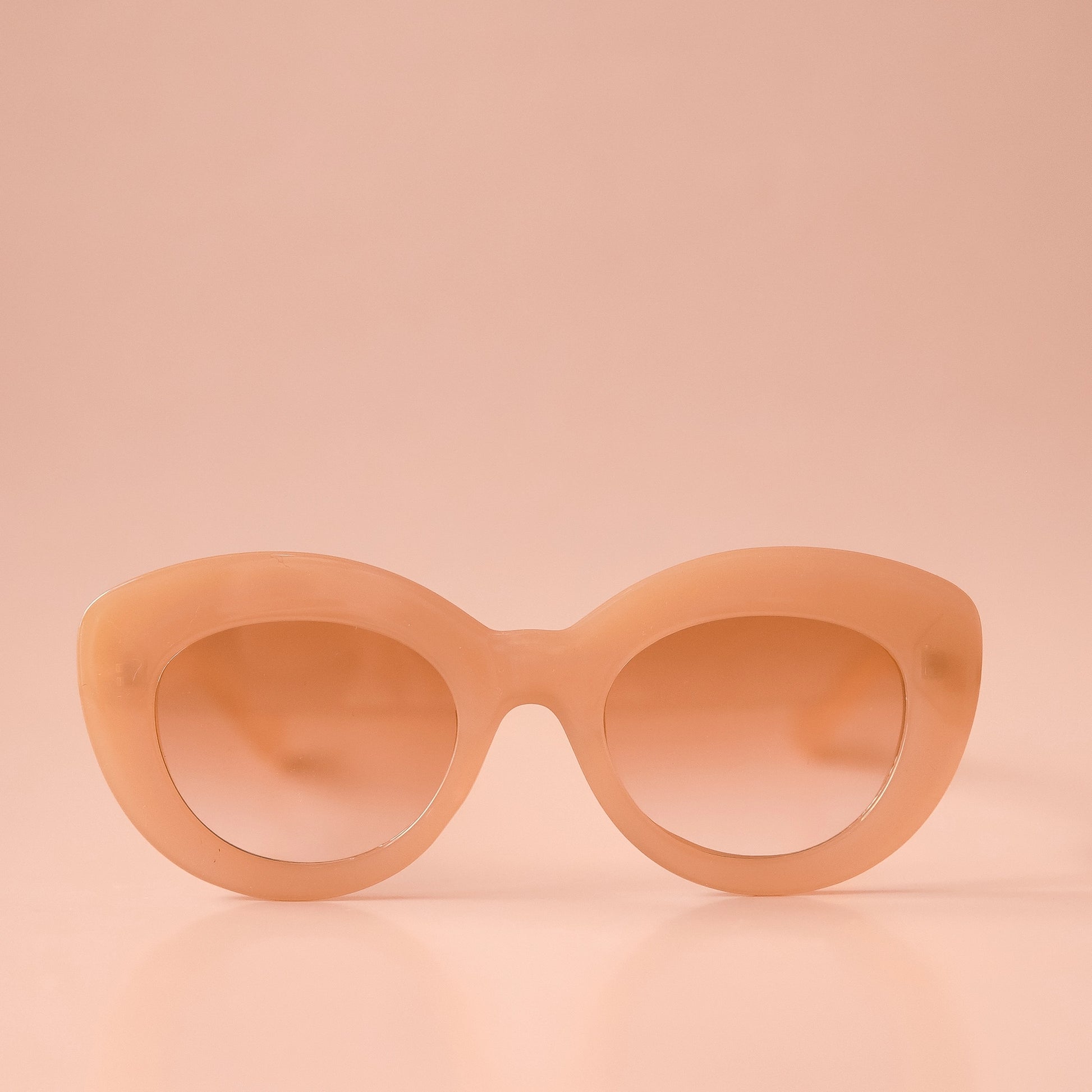 On a peachy background is the Gemma Sunglasses in the shade Amber. They have a rounded shape with a slight cateye flair at the corners. The frame material is a durable amber / peachy colored plastic and the lenses are a similar tone.