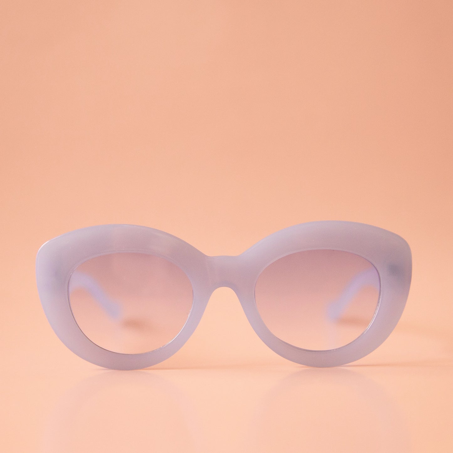 On a peachy background is a pair of light blue sunglasses with a rounded shape and a slight cat eye corner. The lenses are also a light blue shade.