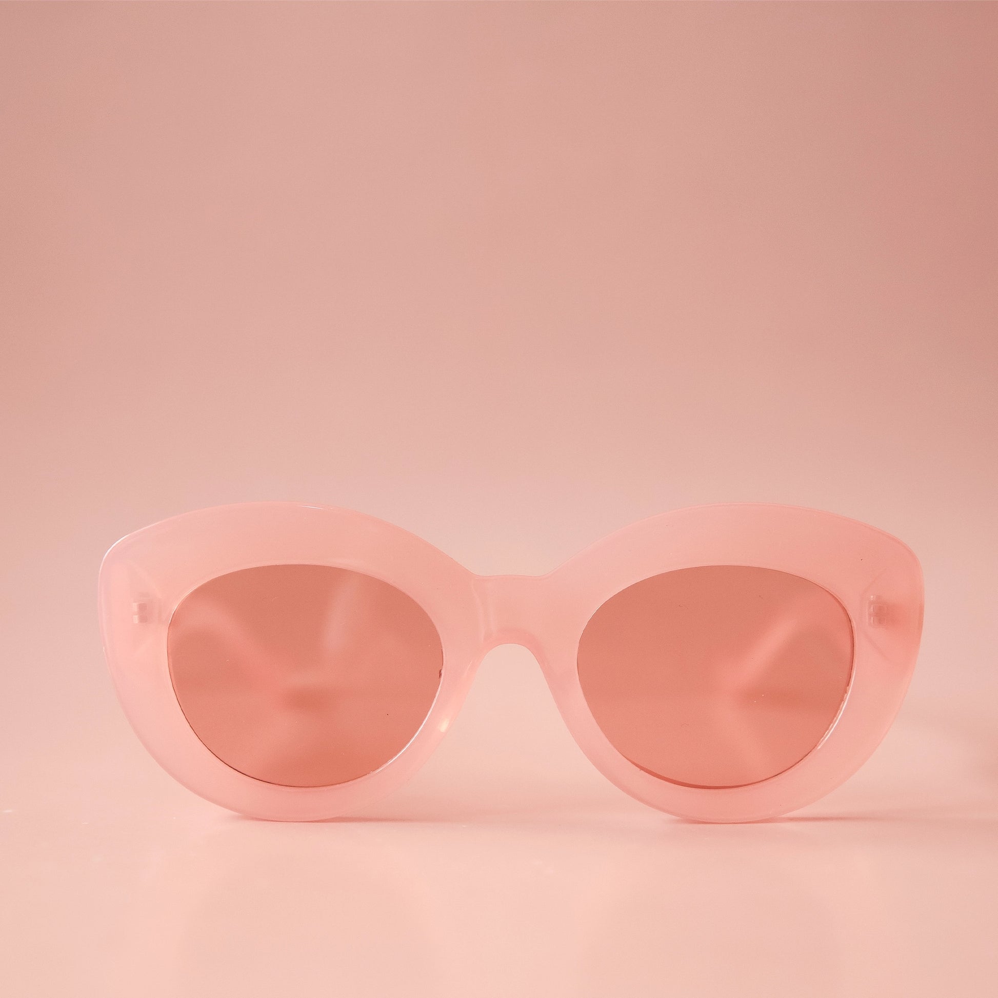 On a light peachy background is a pink pair of sunglasses with a rounded shape and a slight cateye at the corner along with a peachy lens.