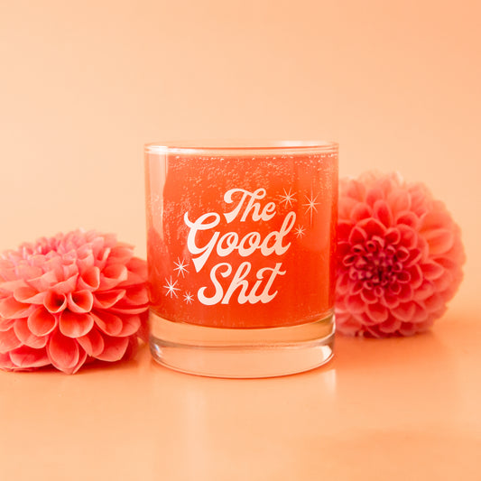 A photograph of a short glass tumbler with a thick bottom and "The Good Shit" printed across the center in white groovy cursive text.