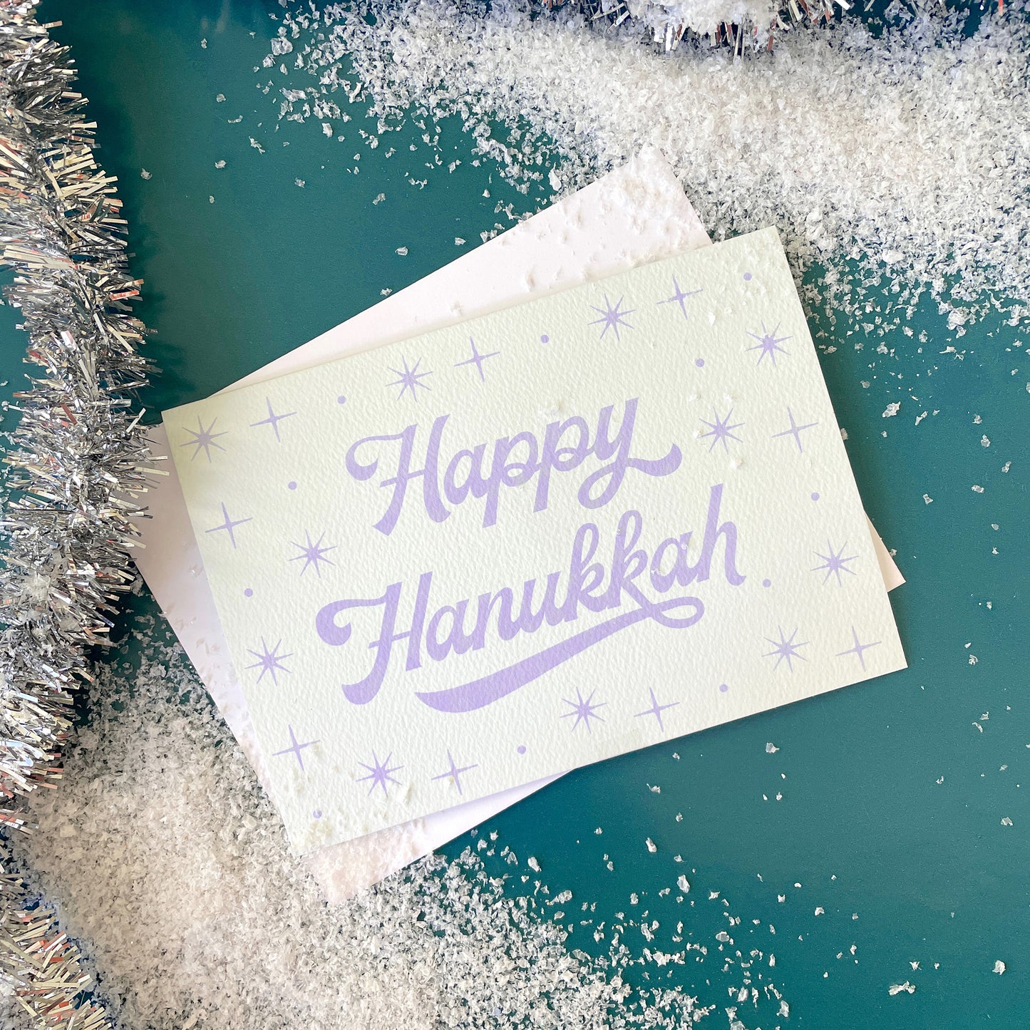 On a teal background is a light blue card that reads, "Happy Hanukkah" with sparkling stars designs around it.