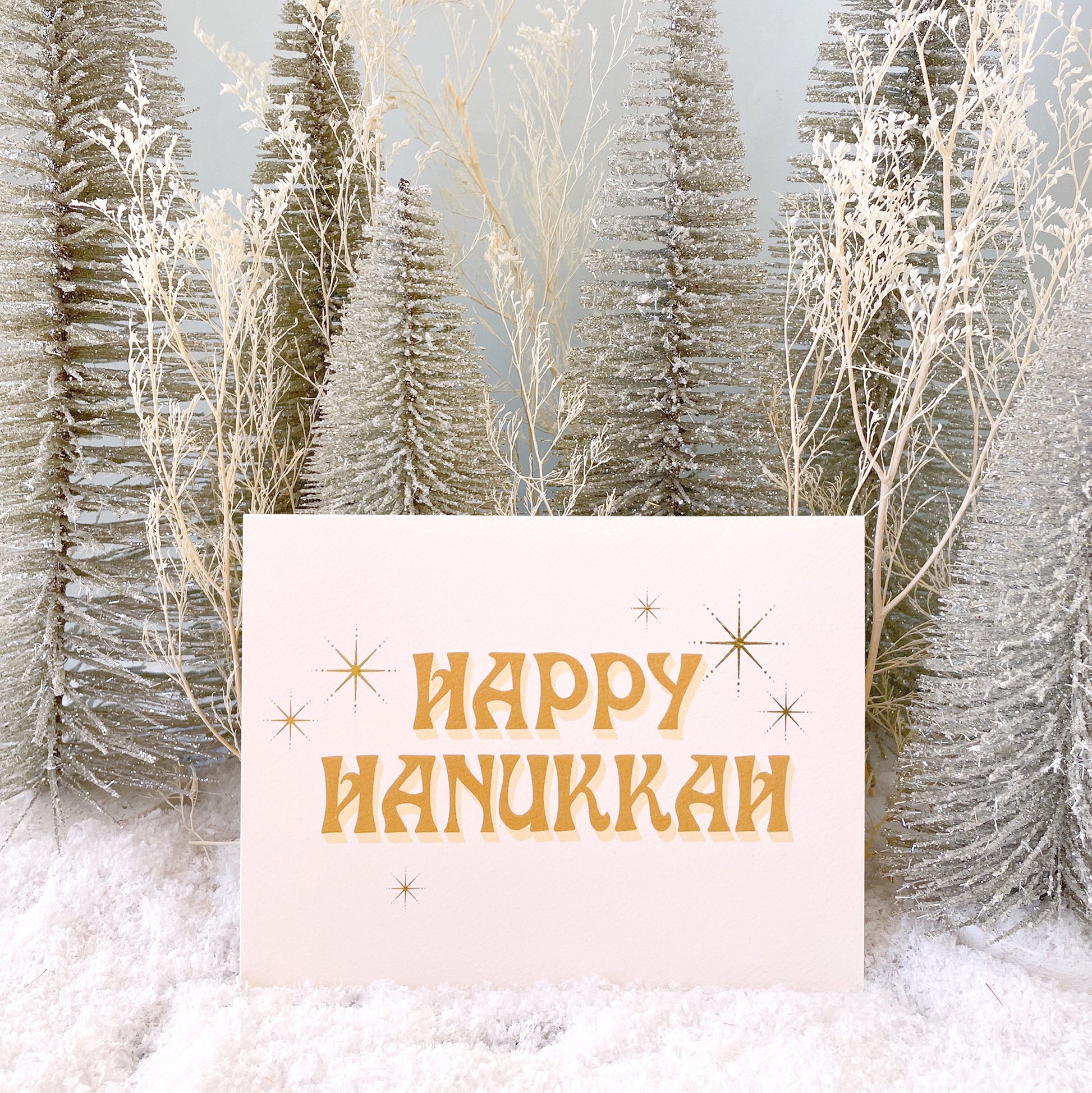 Ivory card that reads 'Happy Hanukkah' in retro orange lettering. The text is accented with gold foil stars twinkling around.