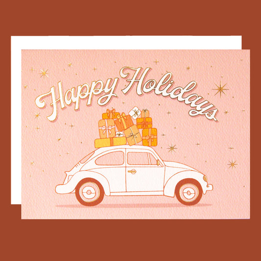 On a pink background is a card with a VW bug with gifts on top and text arched above it that reads, "Happy Holidays".