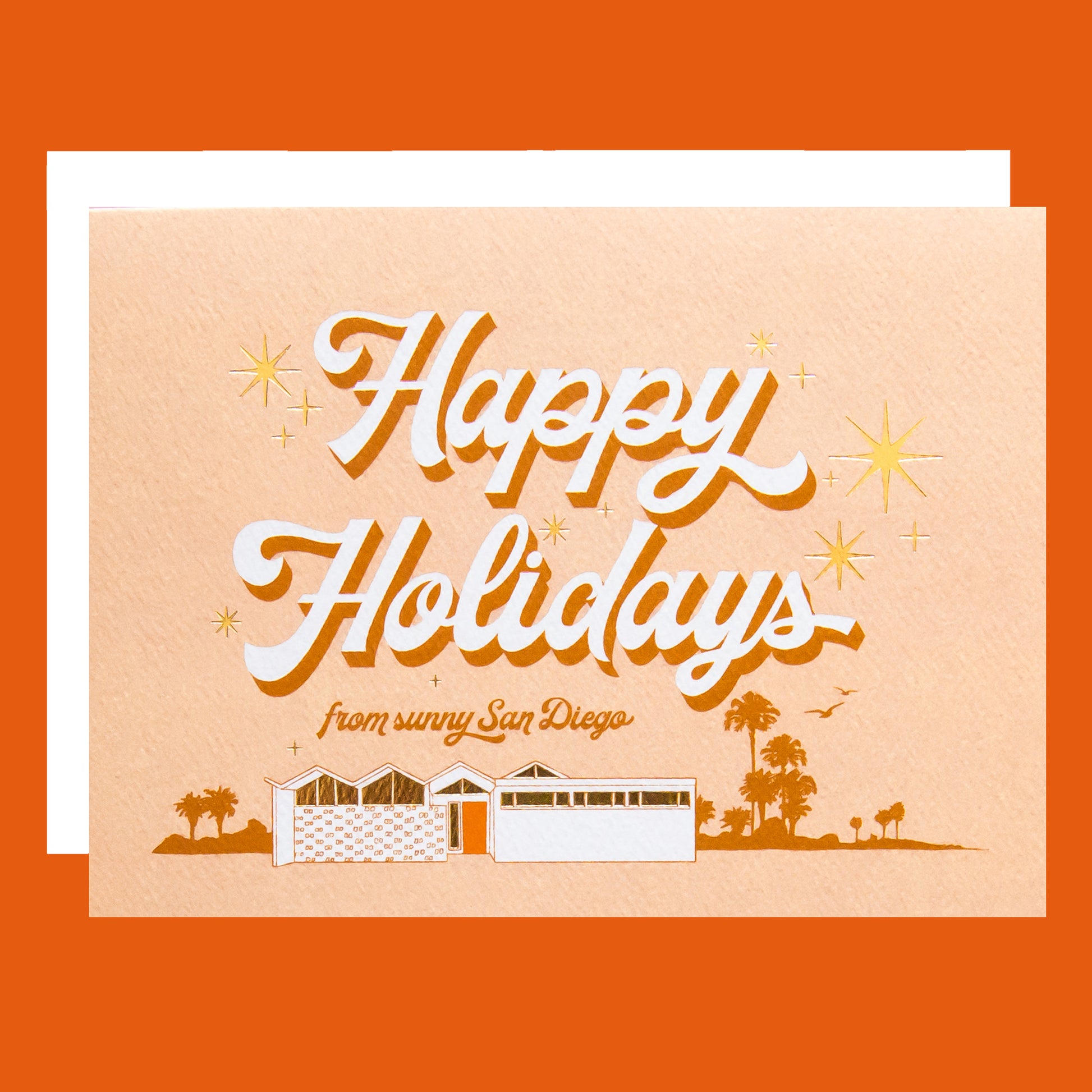 Pale pink card that reads 'Happy Holidays' in white cursive lettering with thin red shadow. Below reads 'from sunny San Diego' in smaller red lettering. Under the text is a coastal scene of a beach house and palm trees. Gold foil sunbursts surround the text. The card is accompanied by a white envelope. 