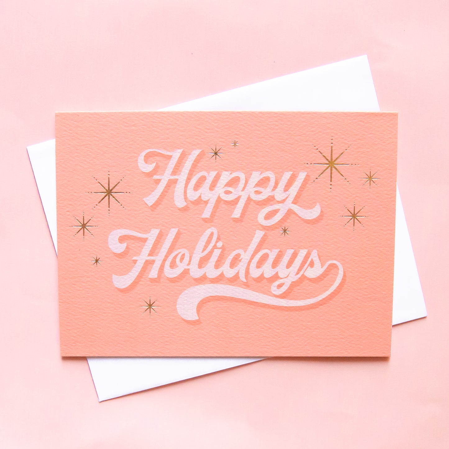 Pink greeting card with white cursive text that reads happy holidays surrounded by six gold foil starbursts. The card is accompanied by a solid white envelope.