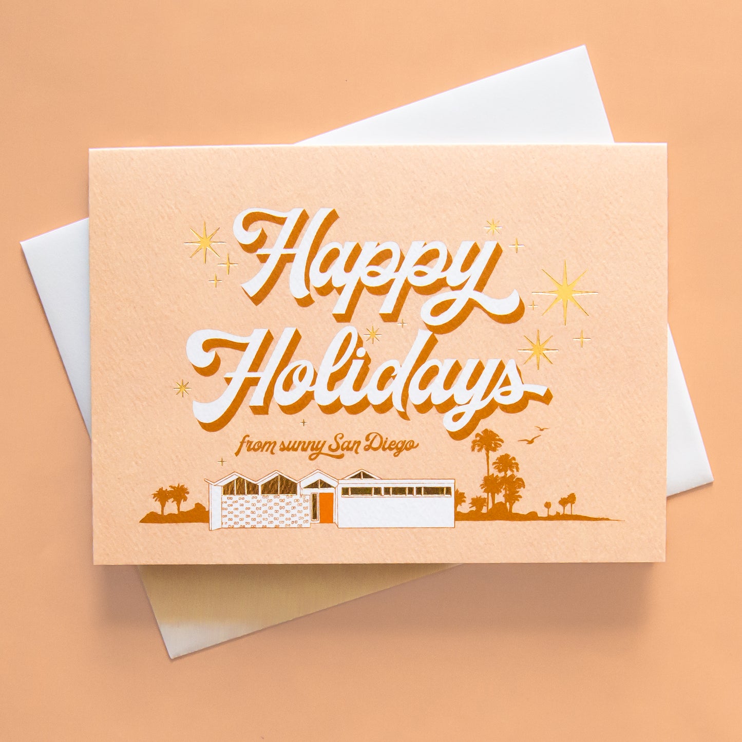 Pale pink card that reads 'Happy Holidays' in white cursive lettering with thin red shadow. Below reads 'from sunny San Diego' in smaller red lettering. Under the text is a coastal scene of a beach house and palm trees. Gold foil sunbursts surround the text. The card is accompanied by a white envelope. 