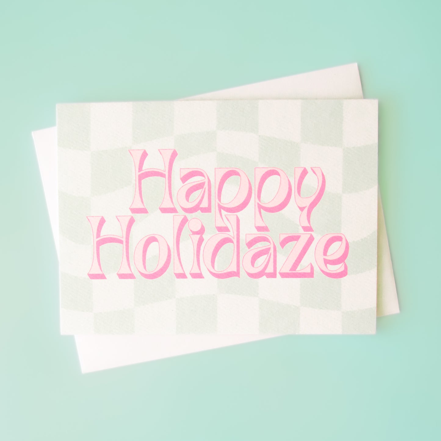 Pale mint and cream checkered card that reads 'Happy Holiday' in retro pink lettering with thin magenta shadow. The card is accompanied by a solid white envelope.