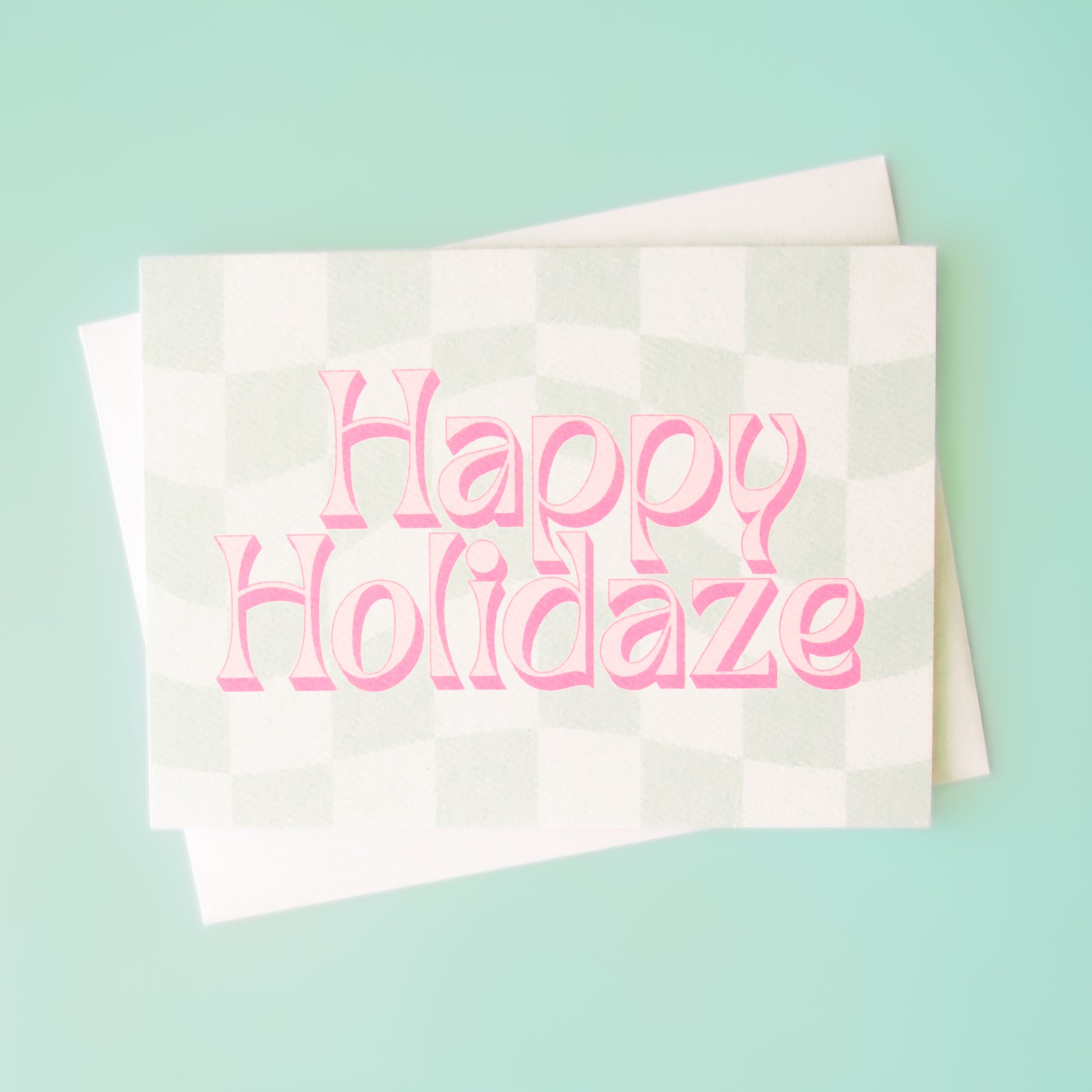 Pale mint and cream checkered card that reads 'Happy Holiday' in retro pink lettering with thin magenta shadow. The card is accompanied by a solid white envelope.