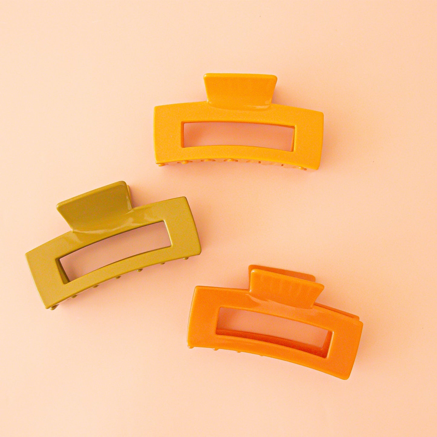On a peachy background is three rectangular shaped claw clips with in three different colors, a mustard yellow, olive green and orange. 
