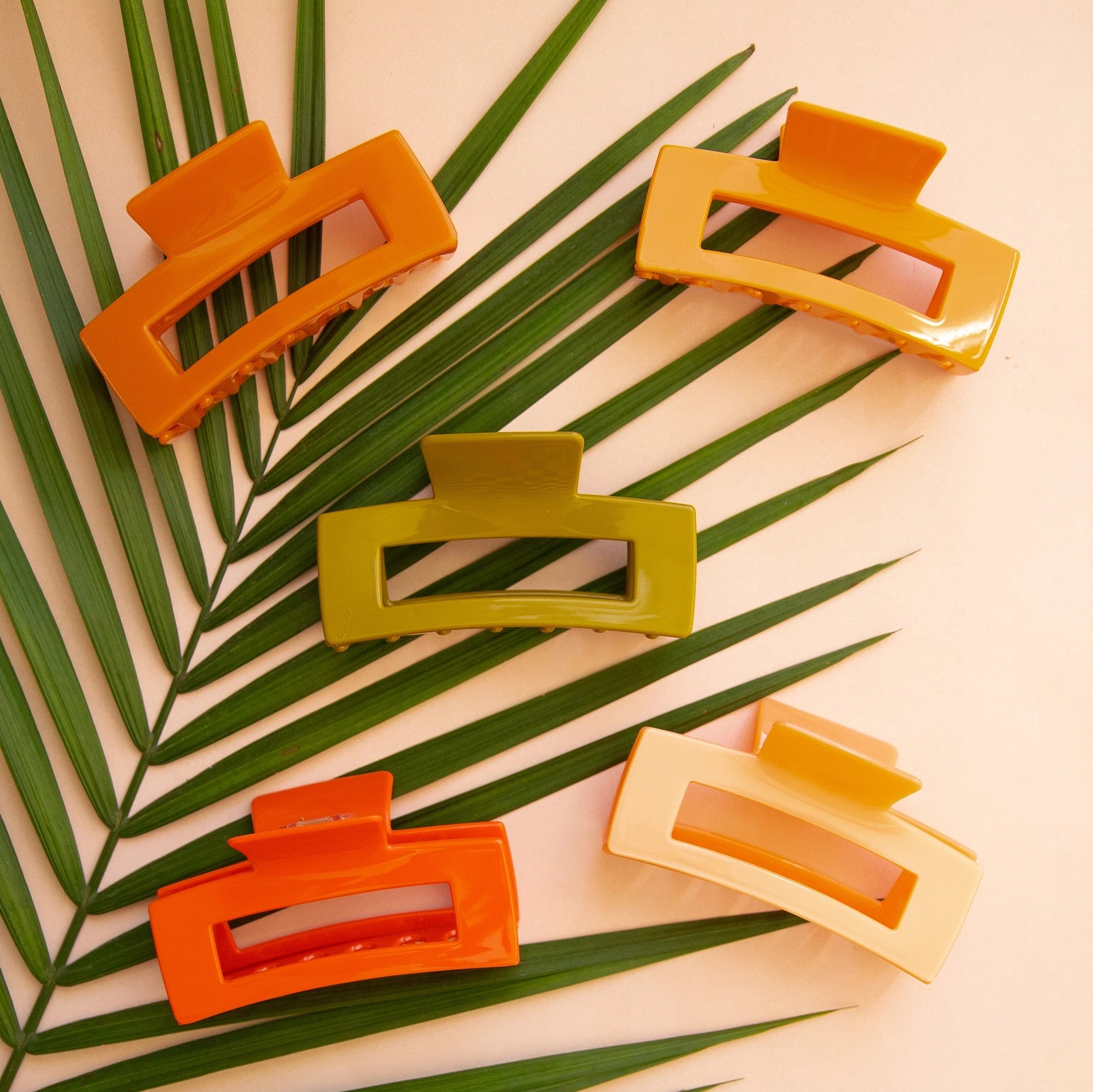 On a peachy background is a variety of rectangle claw clips in different colors on a green palm leaf. 