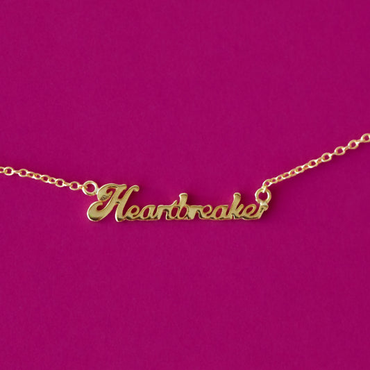 Gold necklace with phrase "heartbreaker" in script on a hot pink background.