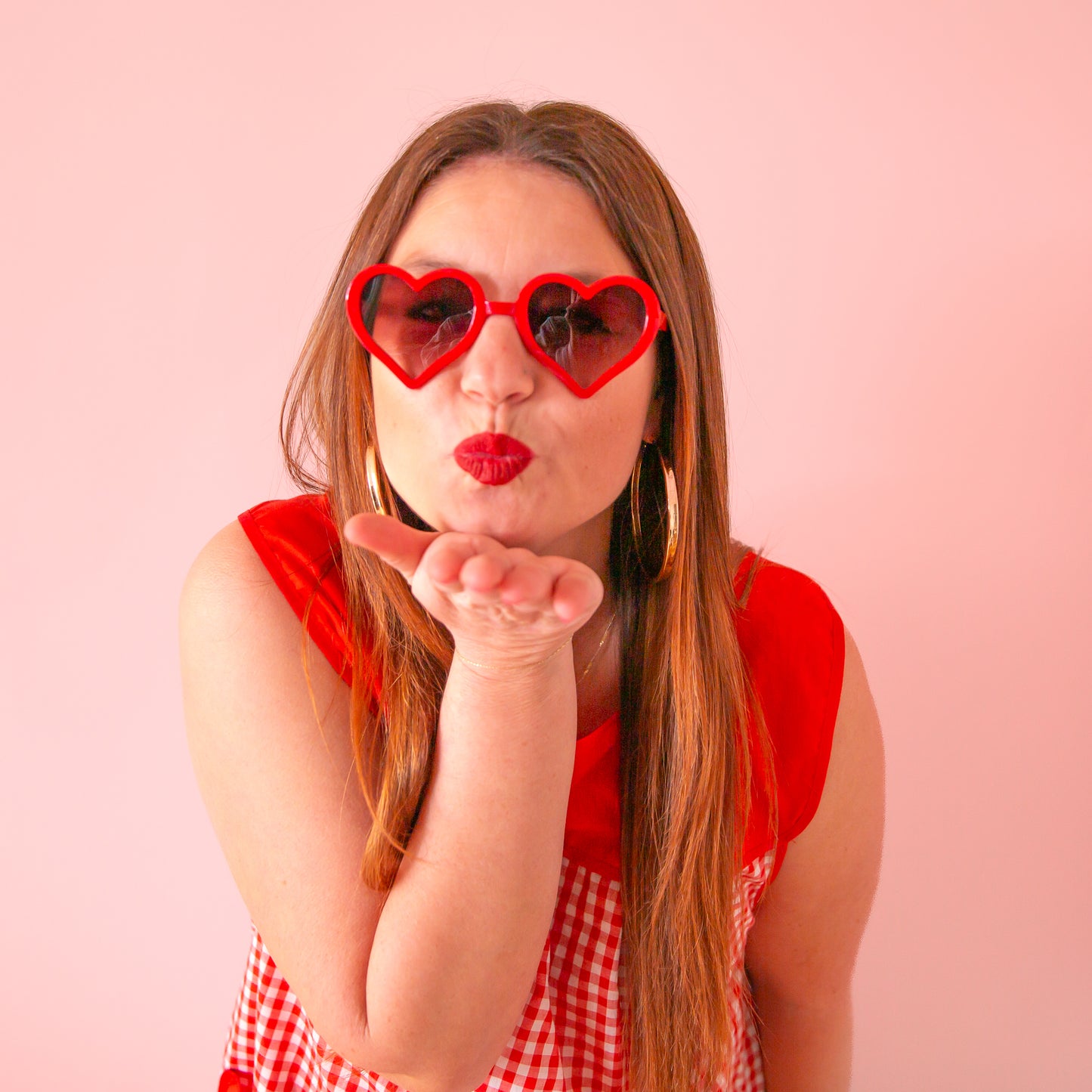 On a pink background is a model wearing the red heart shaped sunglasses.