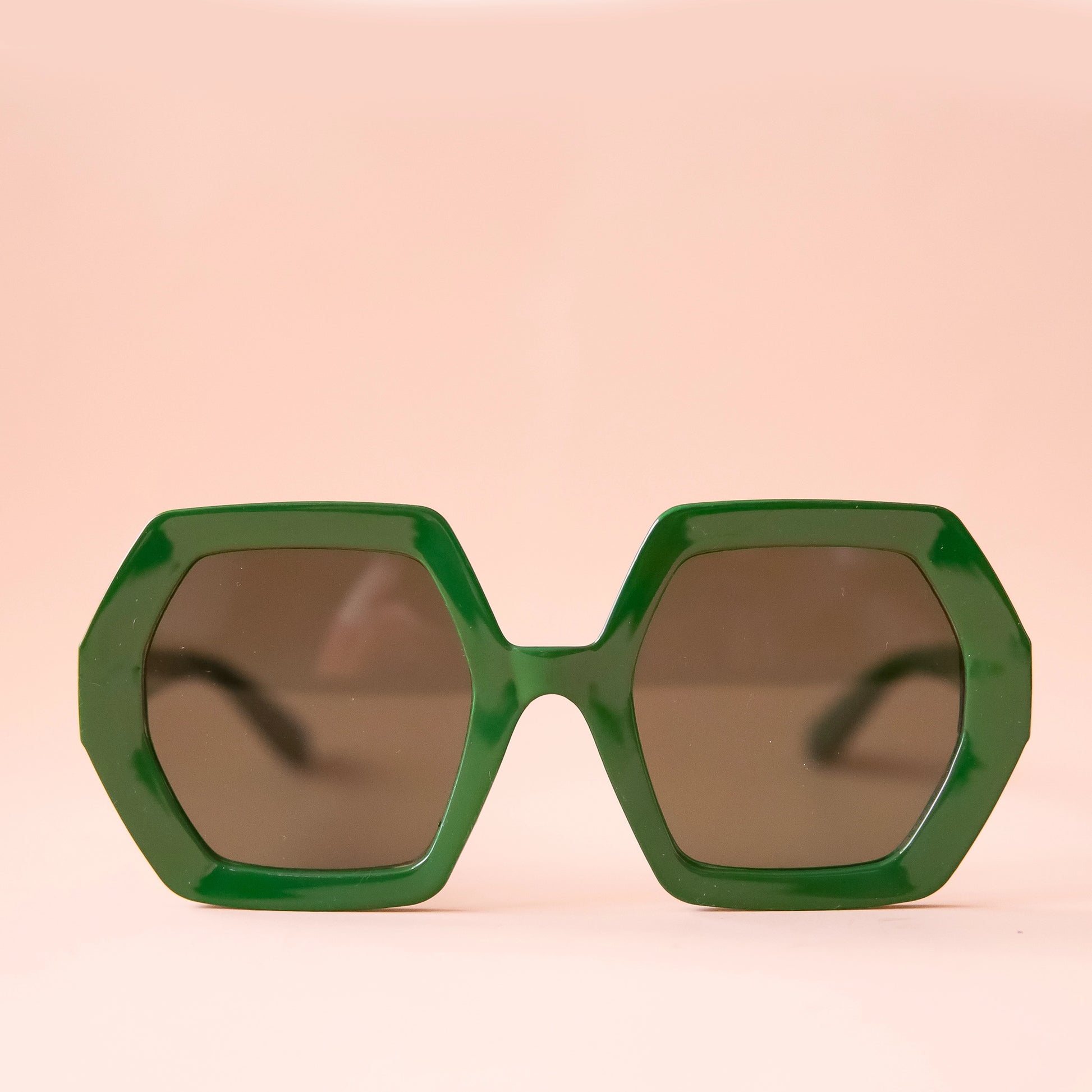 On a peachy background is a pair of hexagon shaped plastic sunglasses in an emerald green color with a dark brown lens.