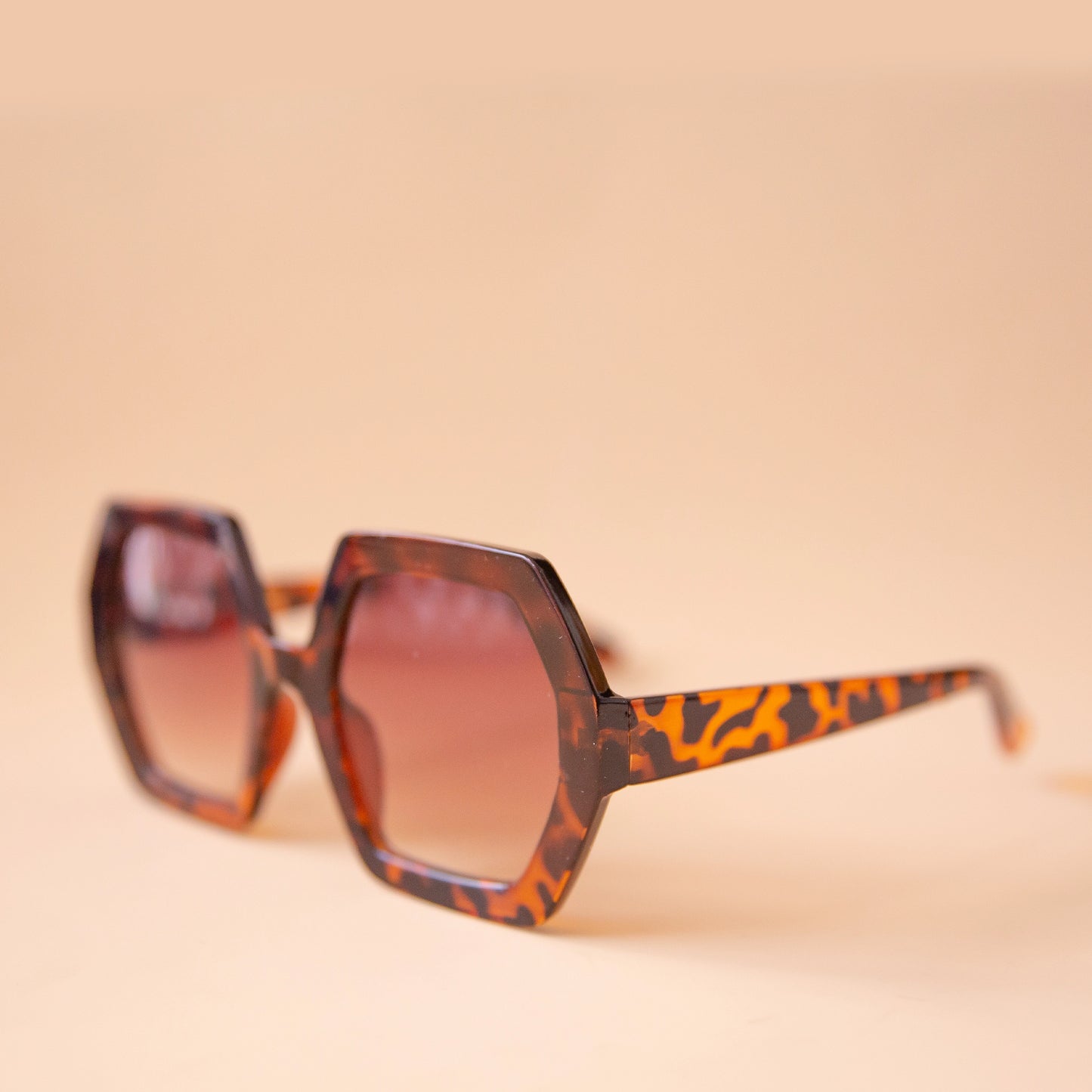 On a peachy background is a pair of hexagon shaped tortoise sunglasses with brown lenses.