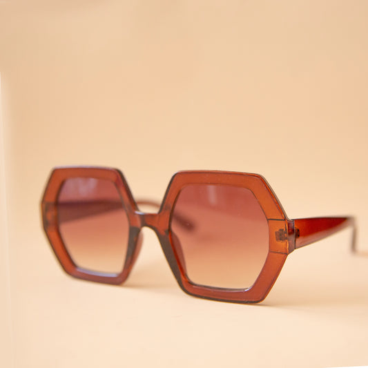 On a peachy background is a pair of hexagon shaped sunglasses in a cognac brown color.