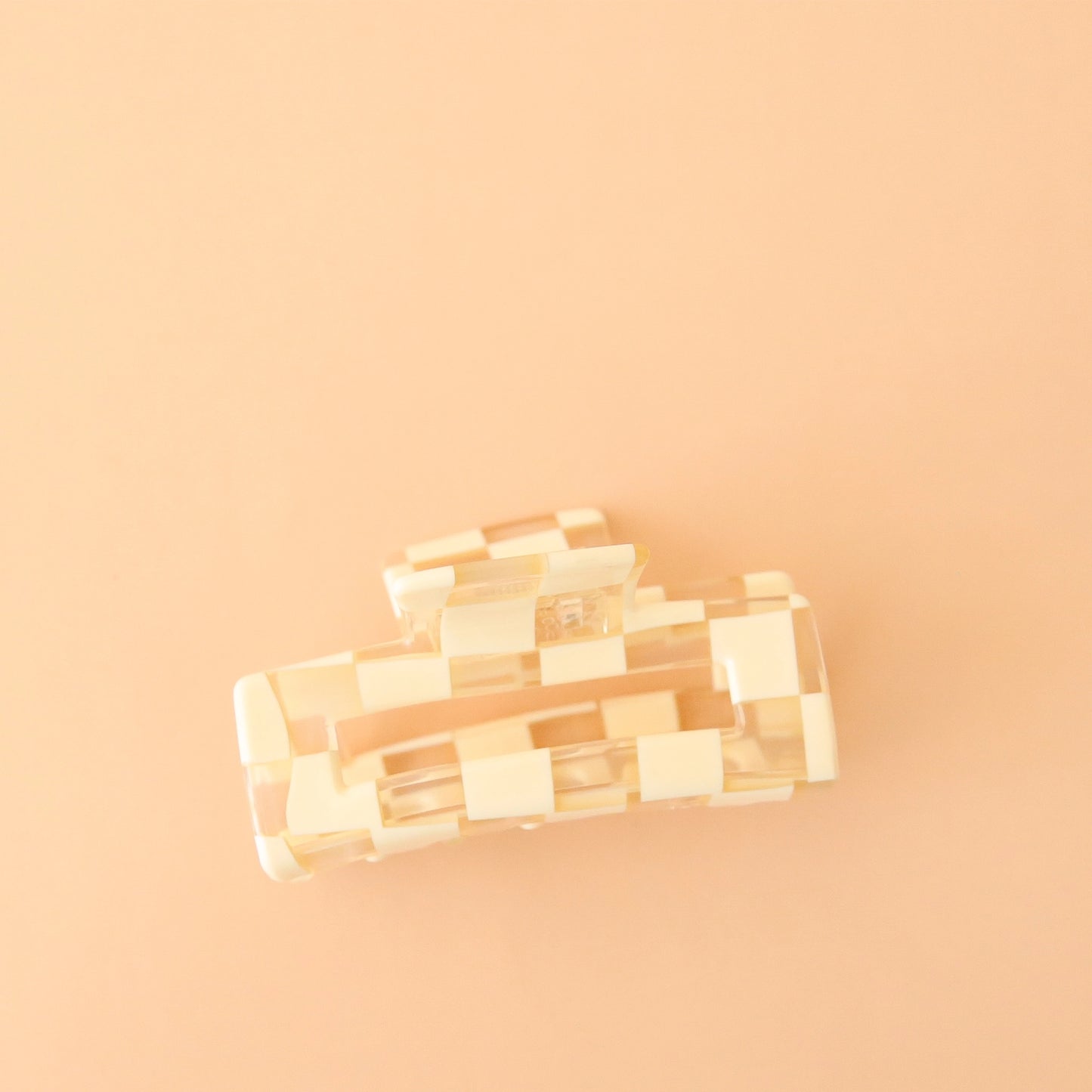 On a peachy background is an ivory and clear checker print claw clip with a rectangle shape. 