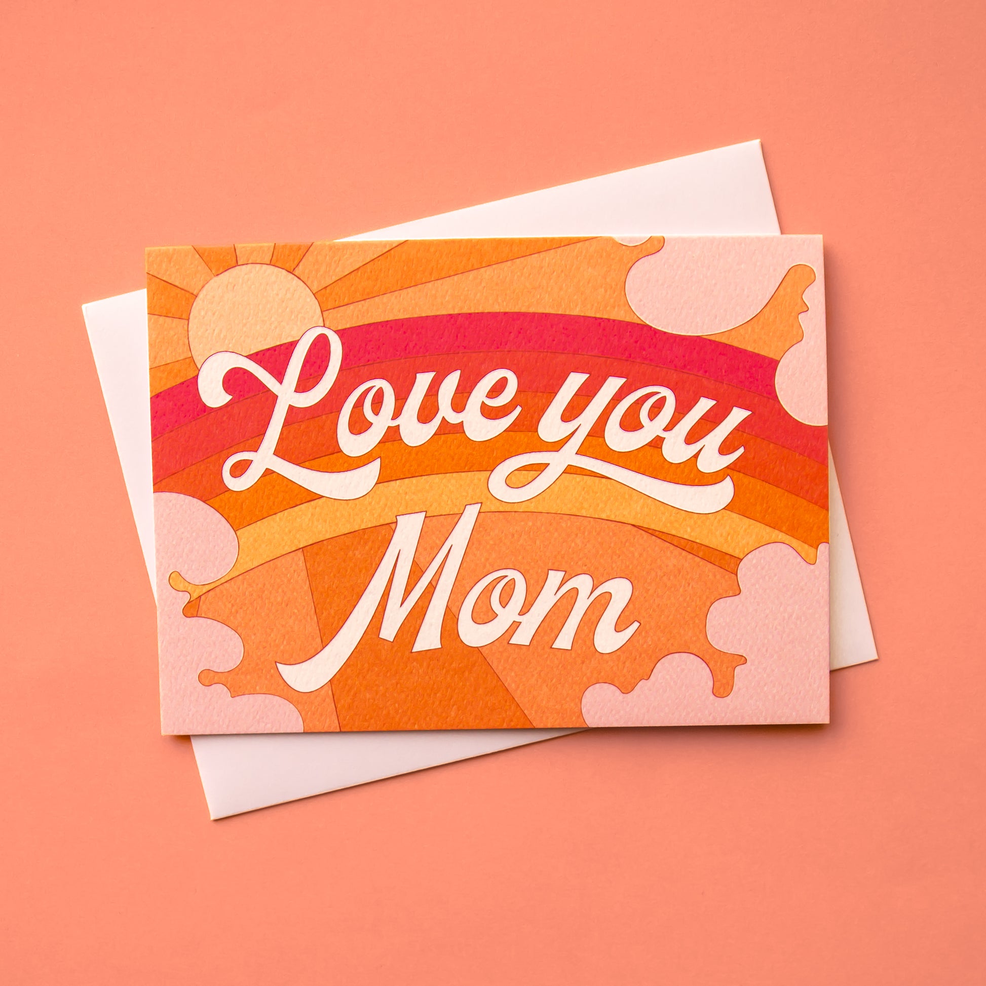 A card with a rainbow graphic made with shades or orange, pink and red along with a shining sun, subtle clouds around the edge and white cursive text that reads, "Love you Mom" in the center as well as a white envelope.