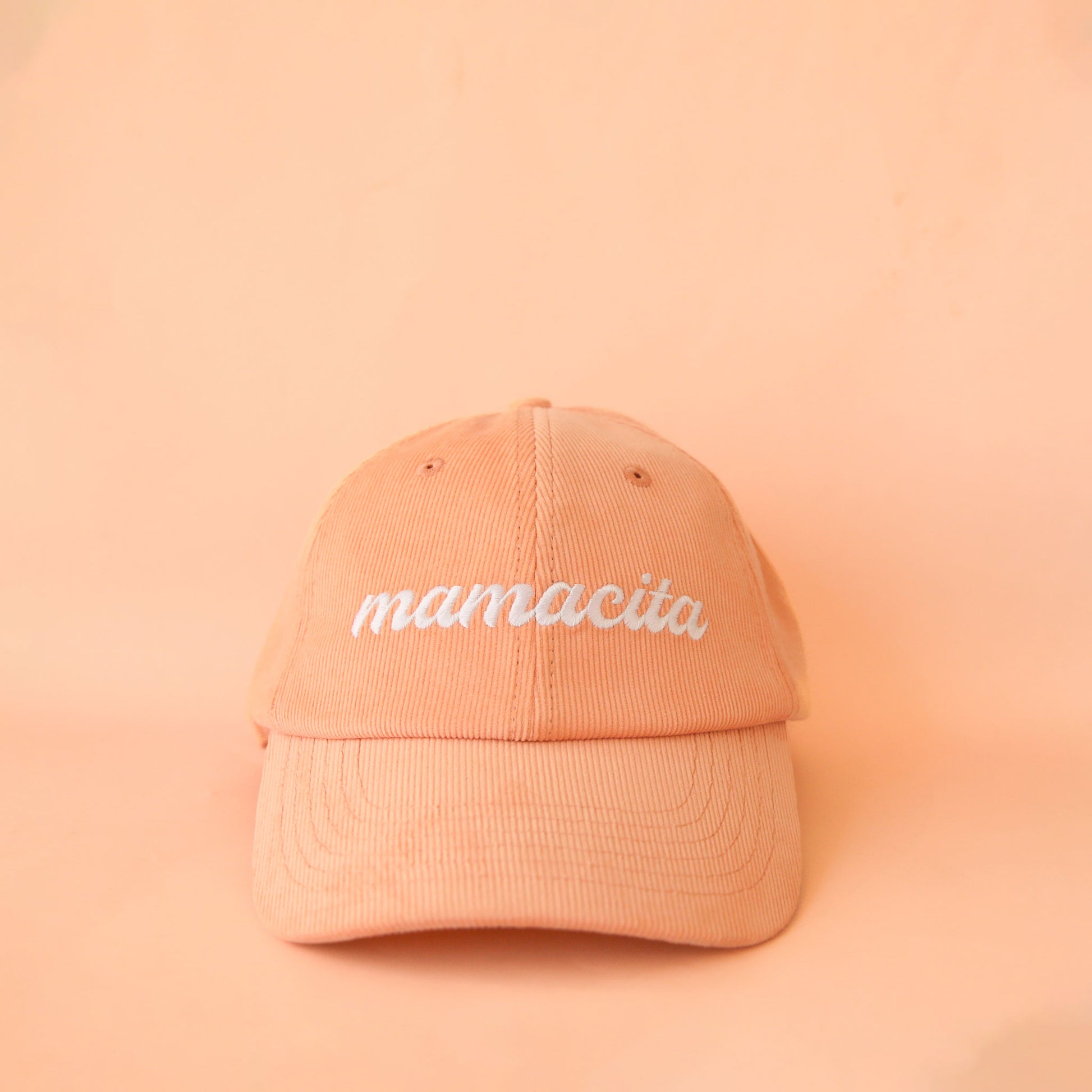 On a peachy background is a peach colored baseball hat with white embroidered text across the front that reads, "mamacita". 