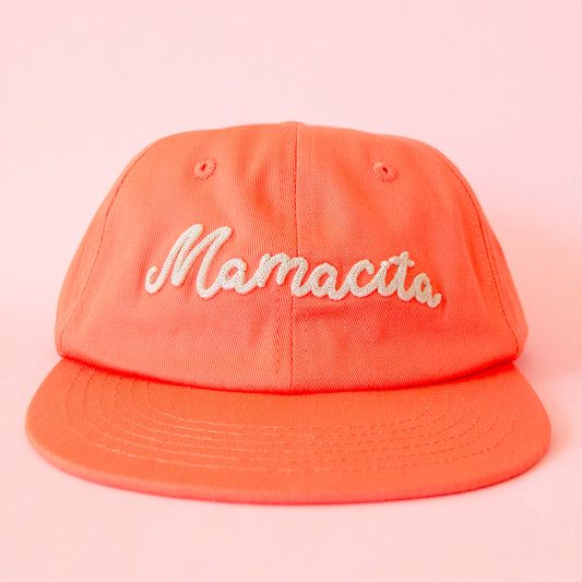 A coral orange flat brim hat with ivory embroidering that reads, 'Mamacita'. 