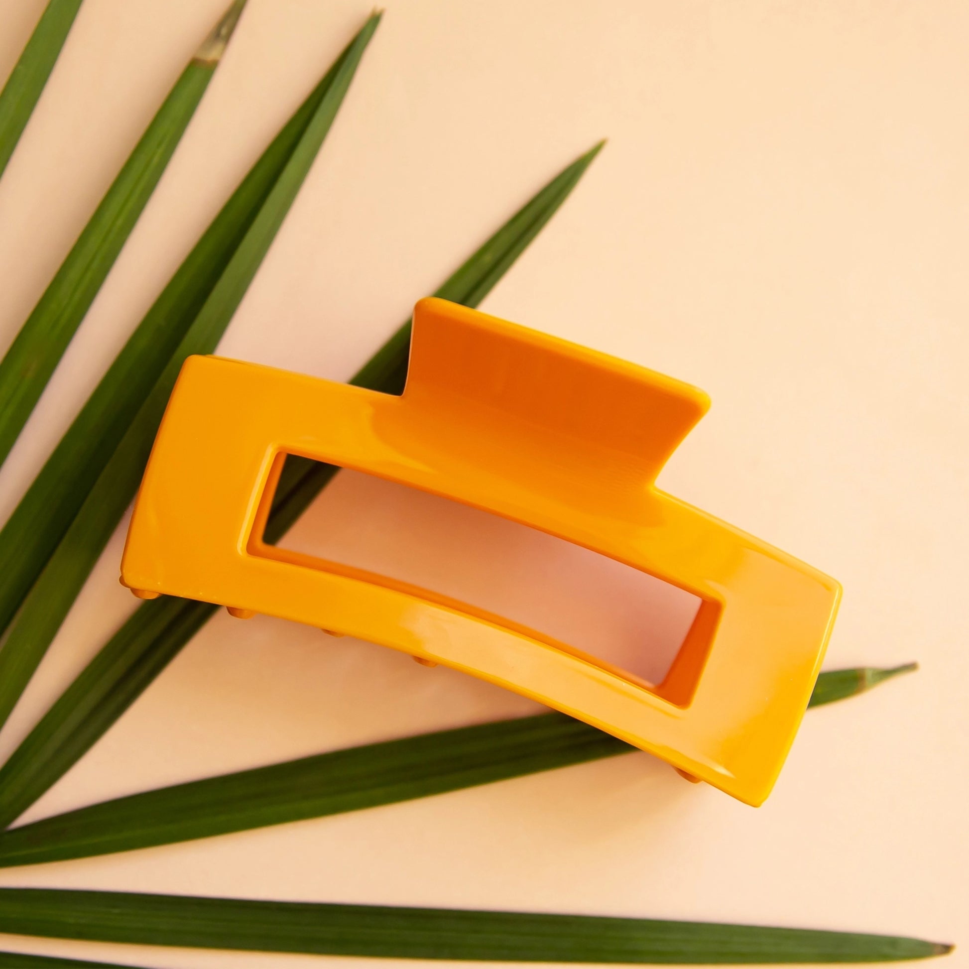 On a peachy background and laying on top of a flat palm leaf is the rectangular claw clip in the shade mango which is a vibrant yellow/orange color.