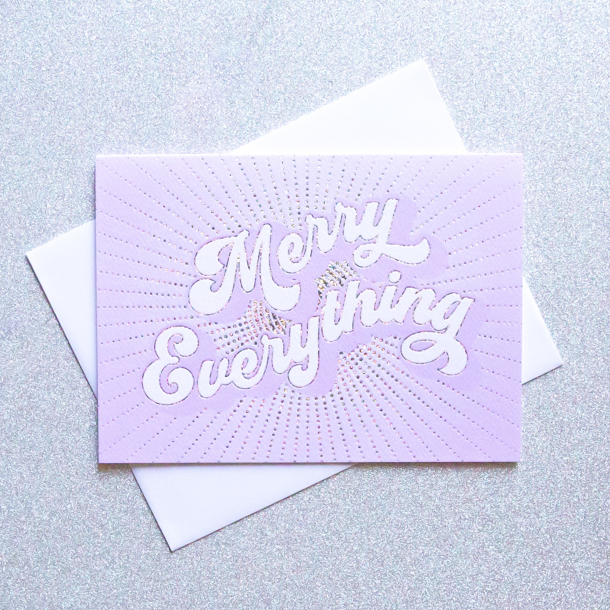 On a sparkle background is a purple greeting card with white text that reads, "Merry Everything" along with a white envelope. 