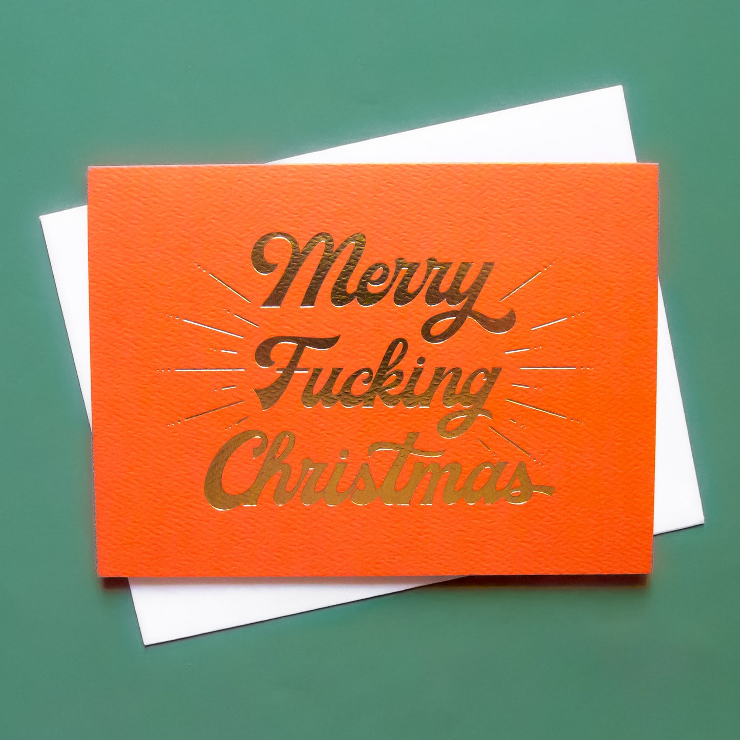 Red card that reads 'Merry Fucking Christmas' in soft pink lettering with dark red shadowing. A gold foil burst detailing beams from the text.