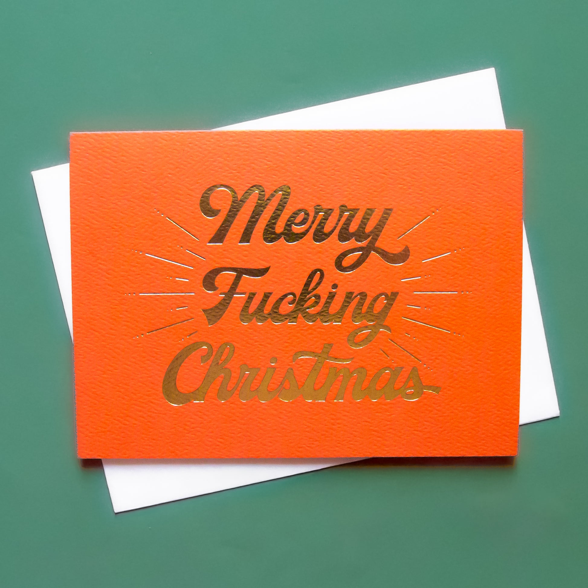 Red card that reads 'Merry Fucking Christmas' in soft pink lettering with dark red shadowing. A gold foil burst detailing beams from the text.