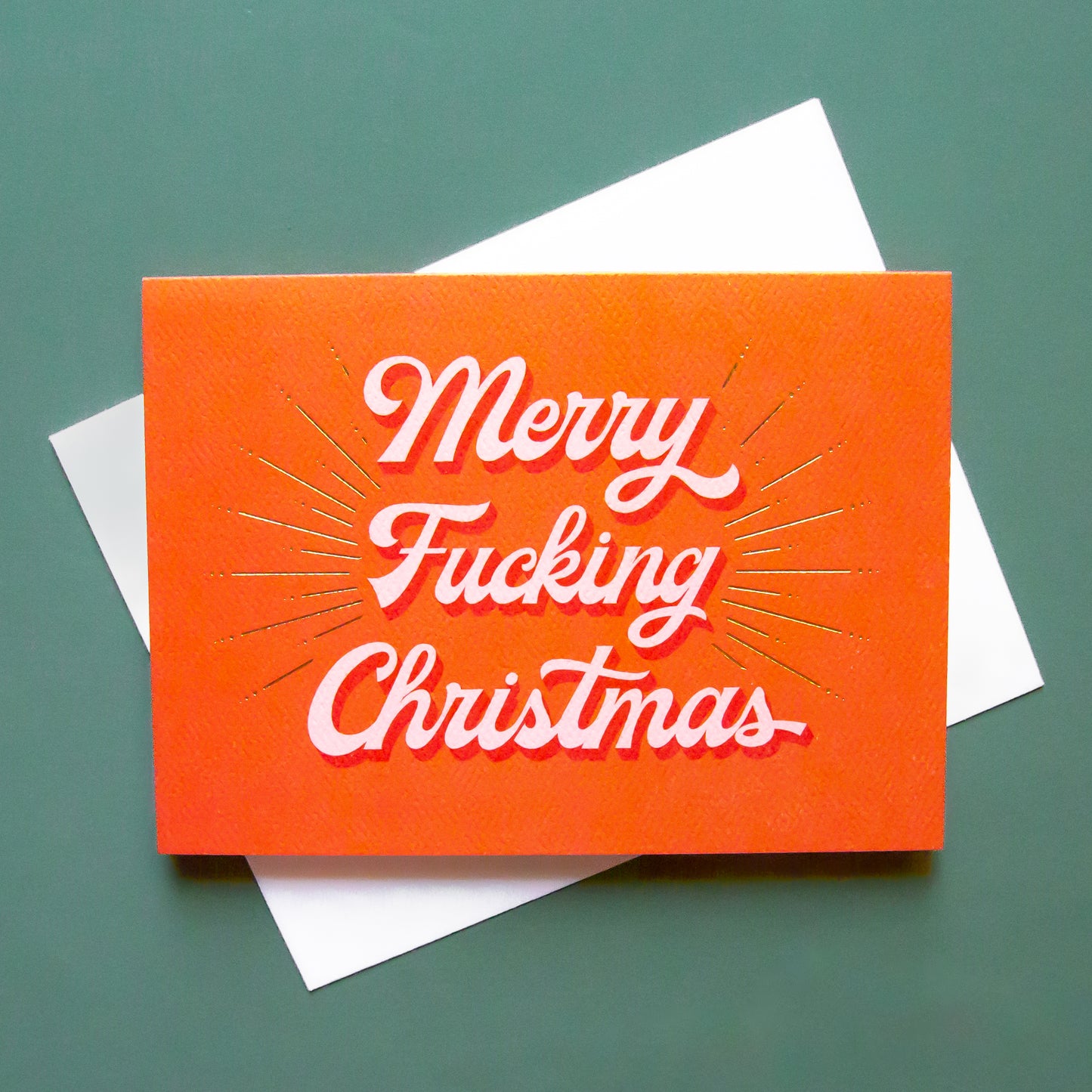 Red card that reads 'Merry Fucking Christmas' in soft pink lettering with dark red shadowing. A gold foil burst detailing beams from the text.