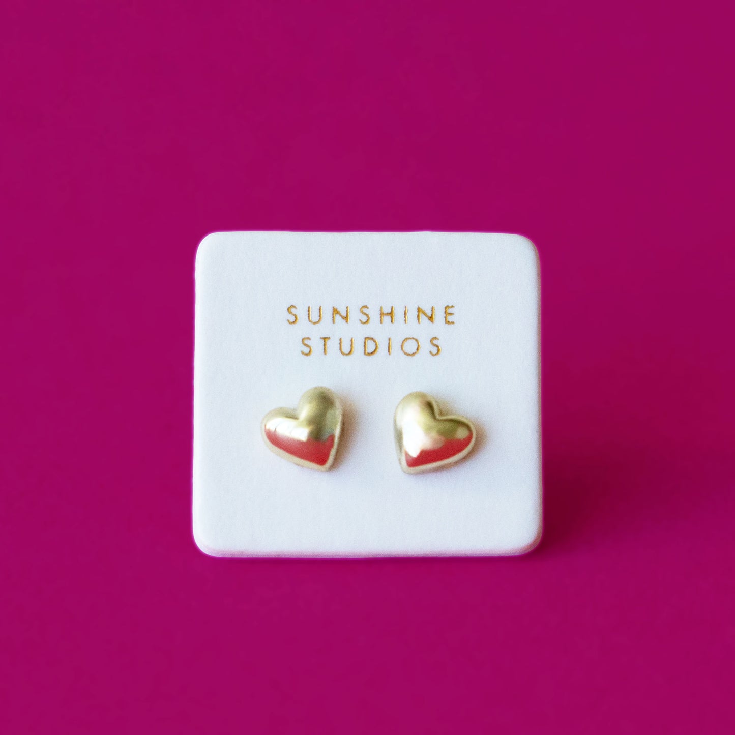 A pair of gold bubble shaped heart earrings on a white backer card and a hot pink background.