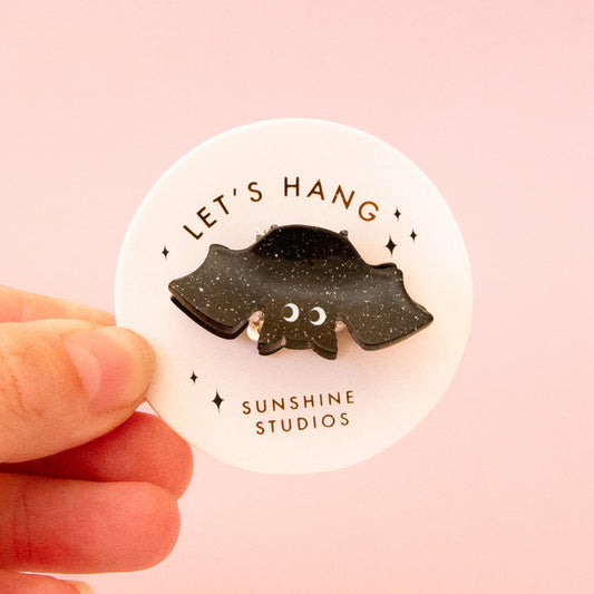 A black speckled bat shaped mini hair clip on a round piece of packaging that reads, 'Let's Hang Sunshine Studios'. 