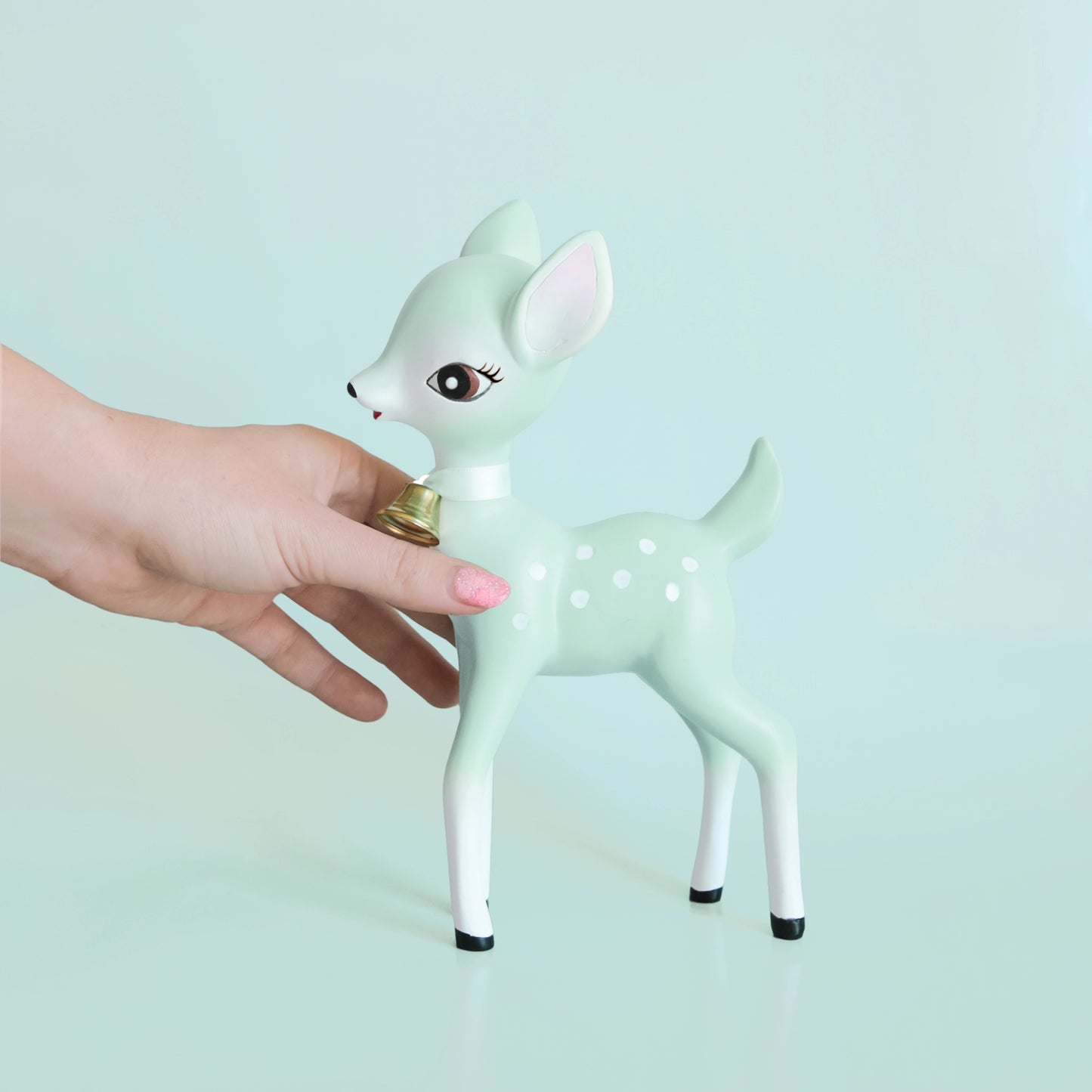 On a light blue background is a mint green retro deer figurine with white spot details and a gold bell at the front of its neck.