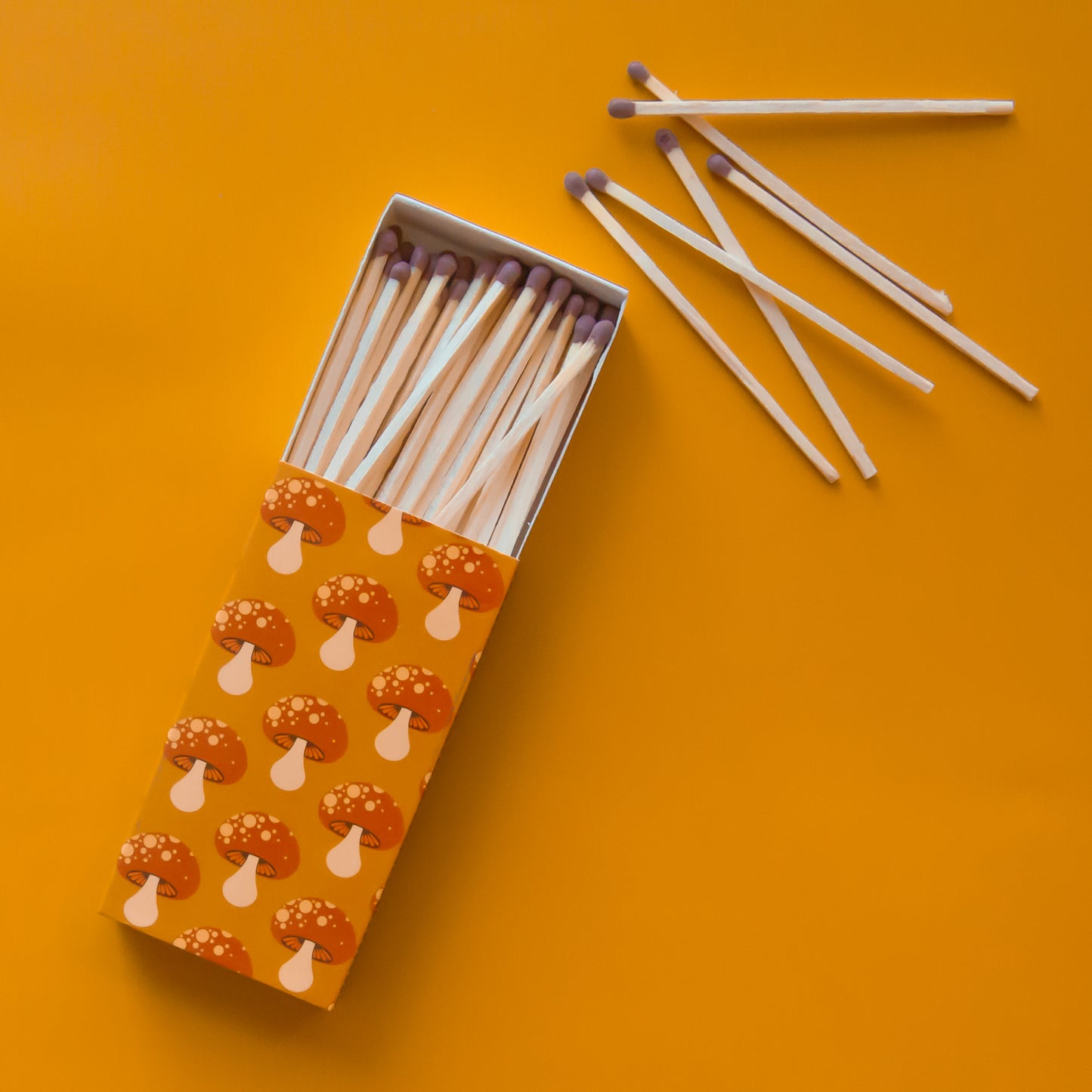A rectangle box of matches with a repeating red mushroom design along all edges besides the top and bottom. The matches that are inside are also photographed here. They are wooden matches the same length as the box with a red striking tip for lighting. 