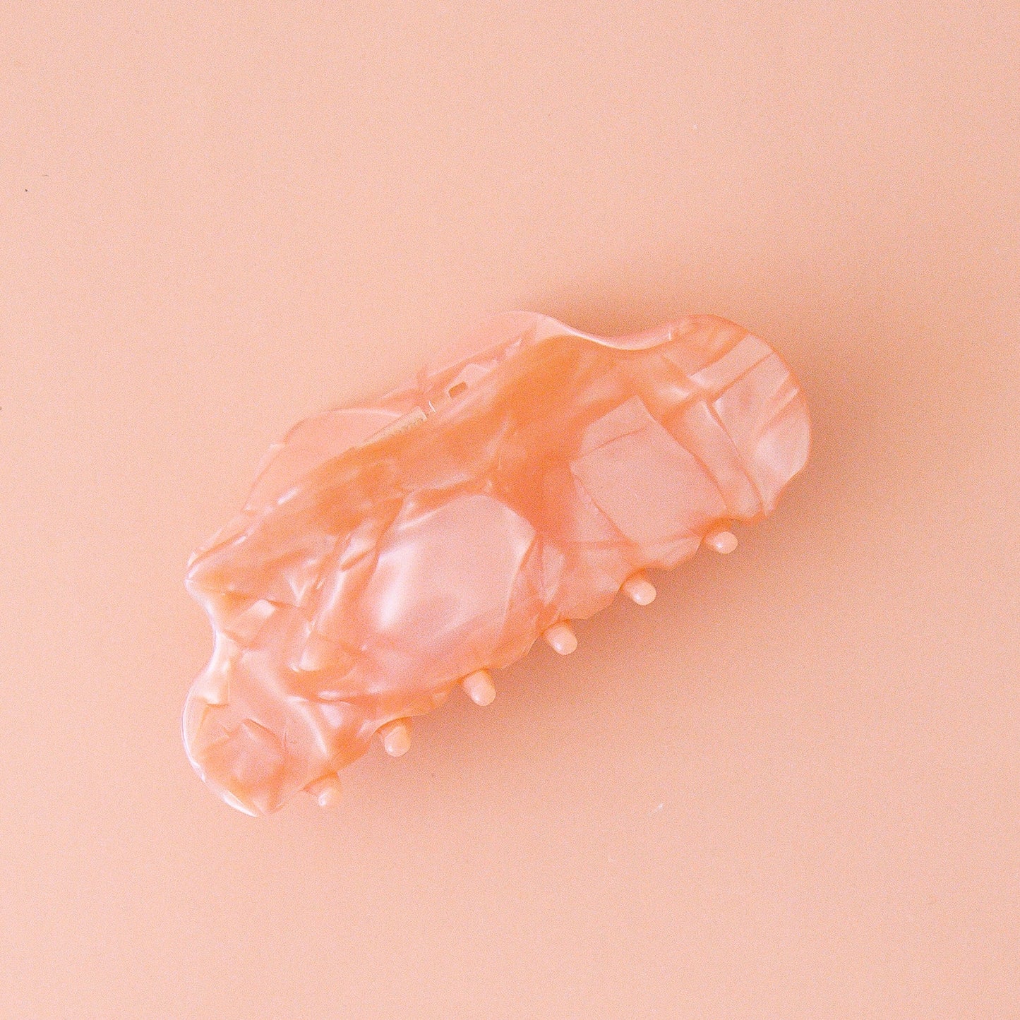 On a light pink background is the venus hair claw clip in the shade peach shell which is a peachy pink color with a shiny marbled finish.