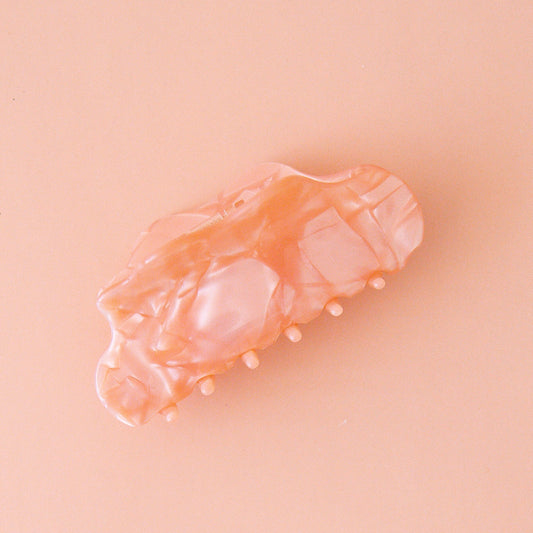 On a light pink background is the venus hair claw clip in the shade peach shell which is a peachy pink color with a shiny marbled finish.