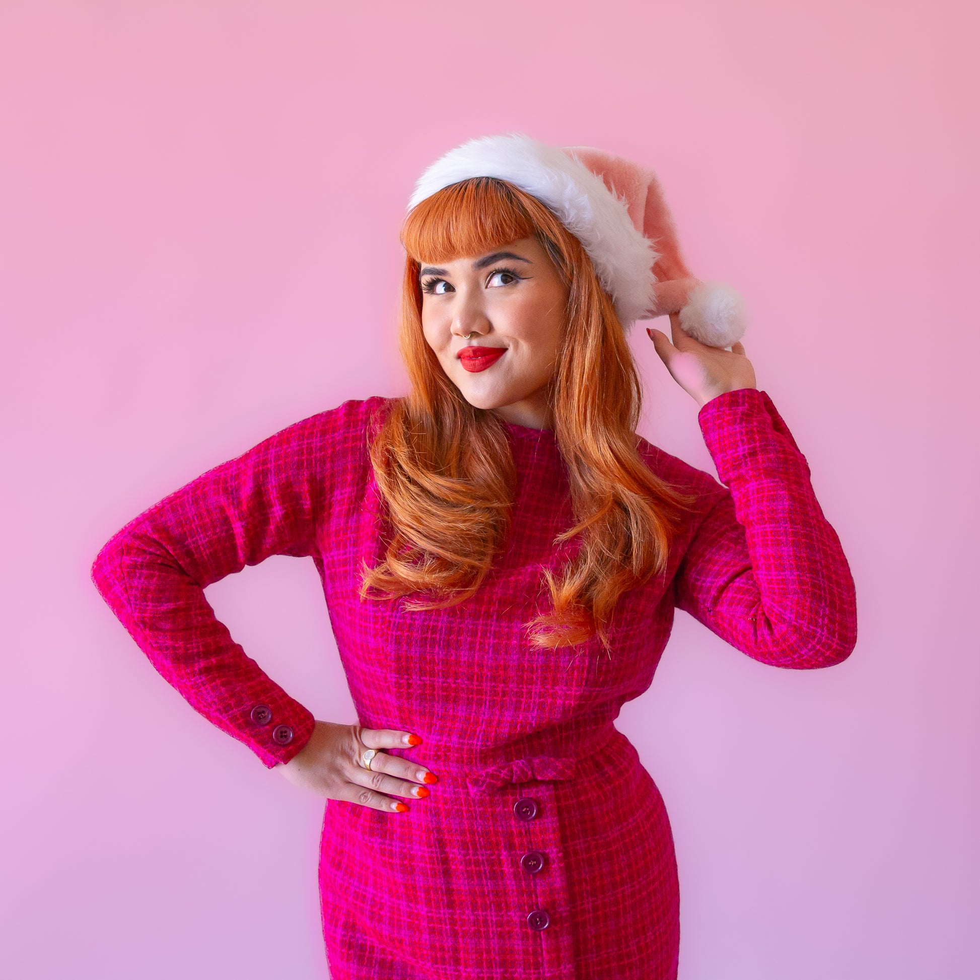 On a pink background is model wearing a pink santa hat with white faux fur detailing.