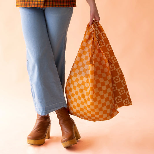 On a peach background is a model holding two nylon reusable bags, one being the playa check which is a dark orange checkered print.
