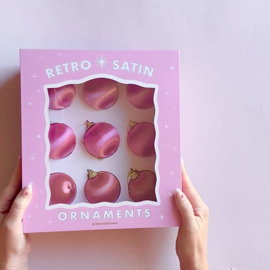 Hands opening a pink box that reads "retro satin ornaments".  Inside the box are 9 round pink and orange hue satin ball ornaments.