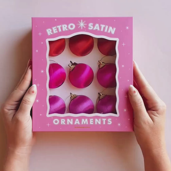 Hands opening a pink box that reads "retro satin ornaments".  Inside the box are 9 round pink and orange hue satin ball ornaments.