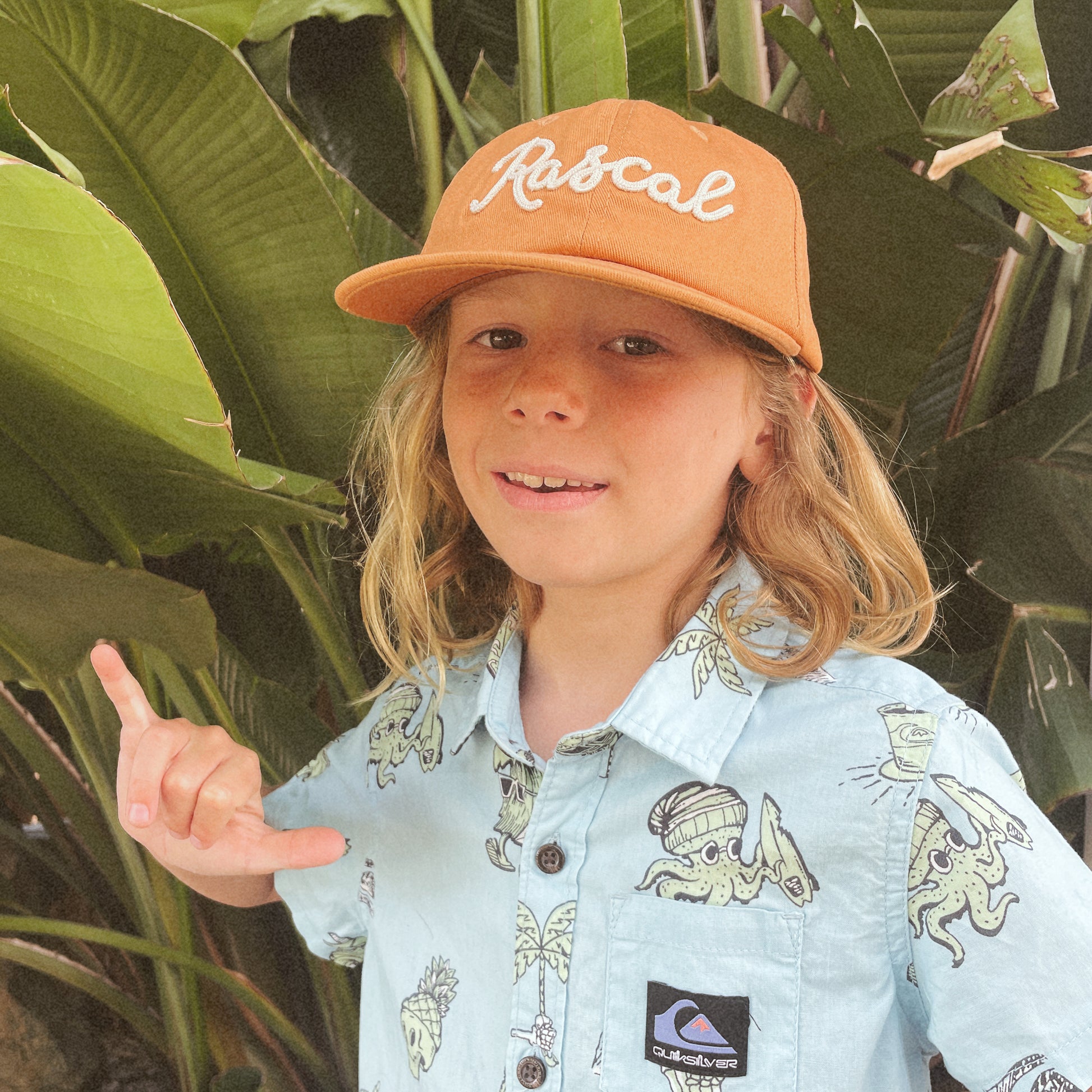 A burnt orange flat brimmed hat with white embroidered text that reads, "Rascal".