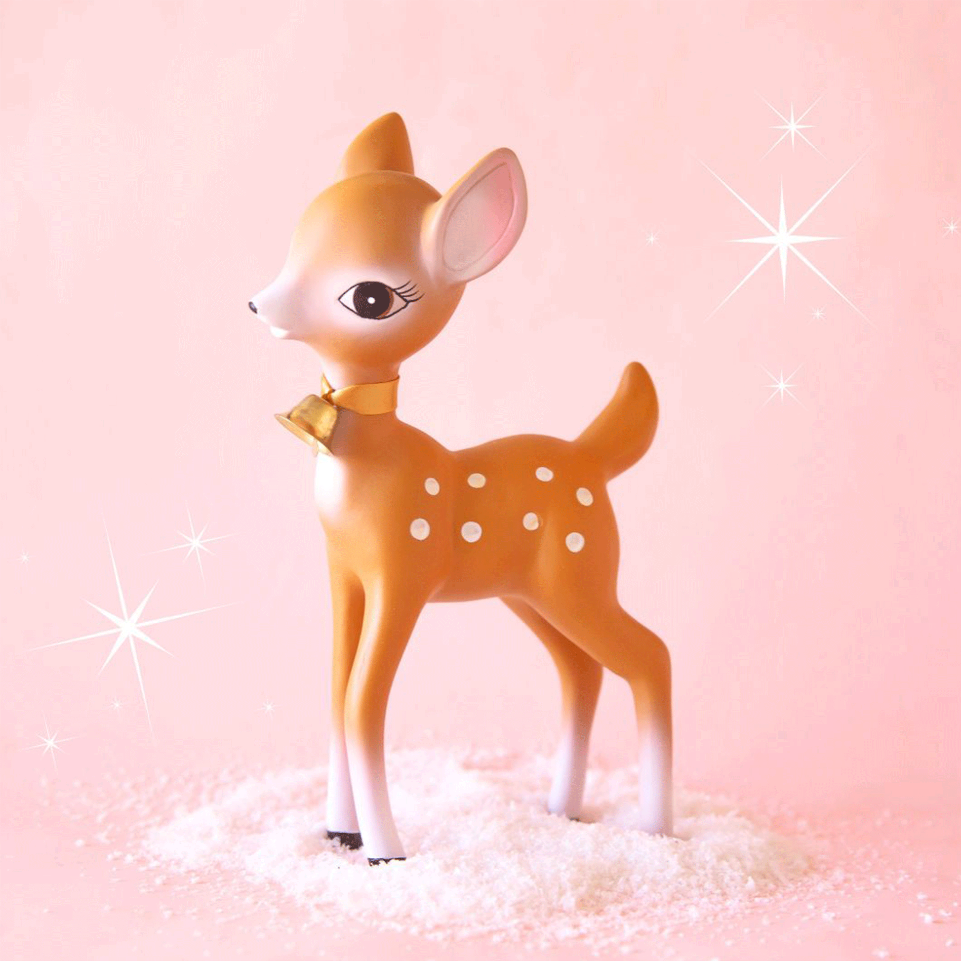 On a peachy background is a brown retro deer figurine with white spots and details, a sweet expression and a gold bell on the front of their neck.