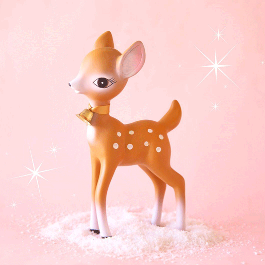 On a peachy background is a brown retro deer figurine with white spots and details, a sweet expression and a gold bell on the front of their neck.