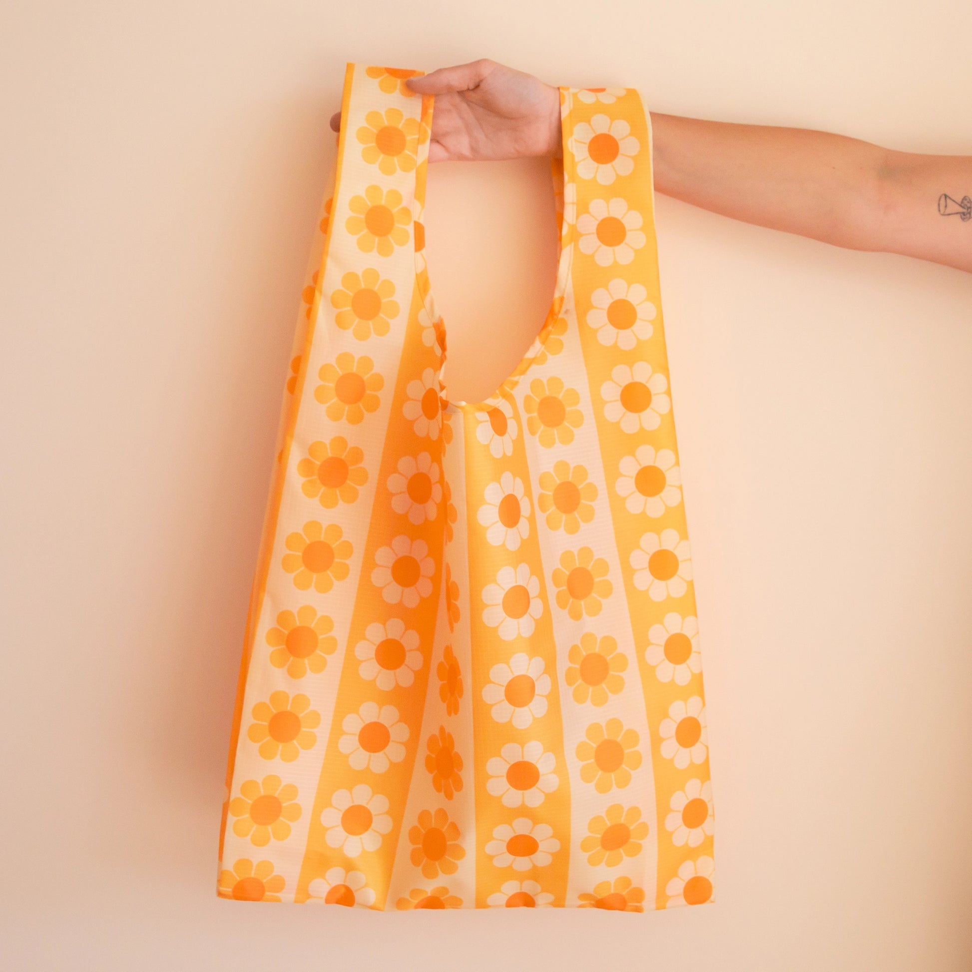 On a tan background if a model's hand holding out a reusable nylon bag with a yellow and orange daisy pattern.  